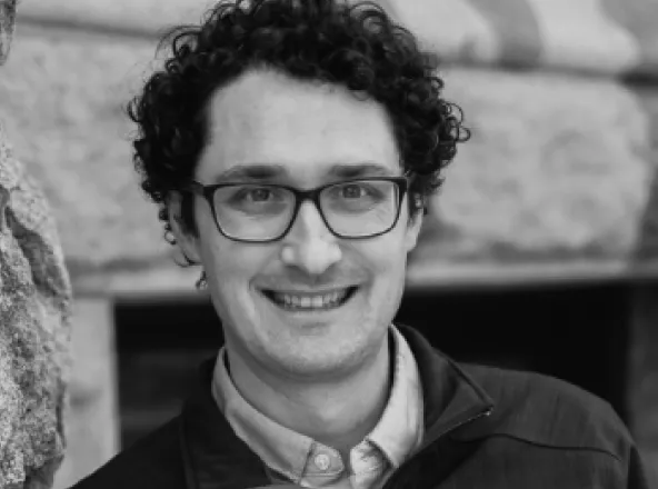 Smiling man with curly hair and glasses wearing a jacket in front of a stone wall.