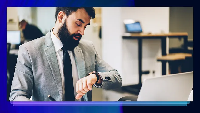 Bearded man in a gray suit looking at his watch in an office setting with a laptop nearby.