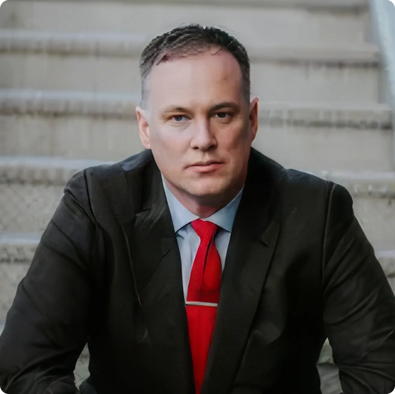 Headshot of an attorney in a dark suit and red tie seated on courthouse steps.