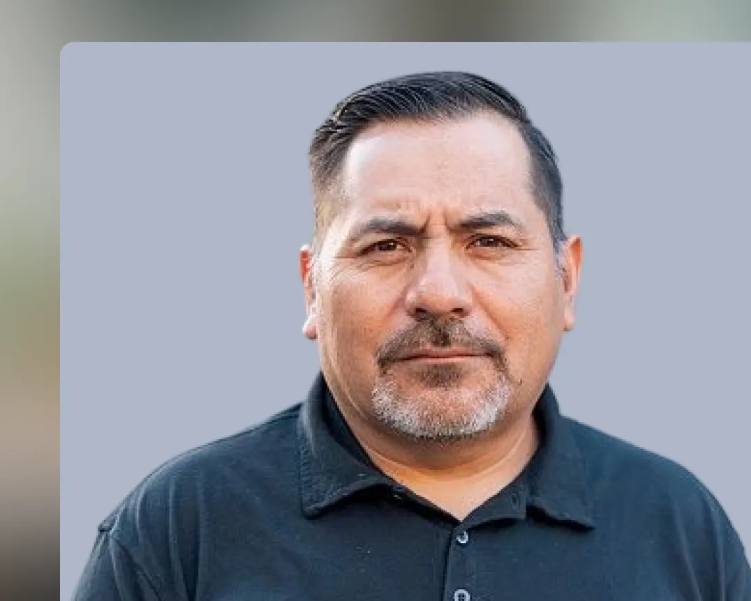 Portrait of a middle-aged man with short dark hair and a trimmed mustache and goatee, wearing a black polo shirt, against a plain light gray background.