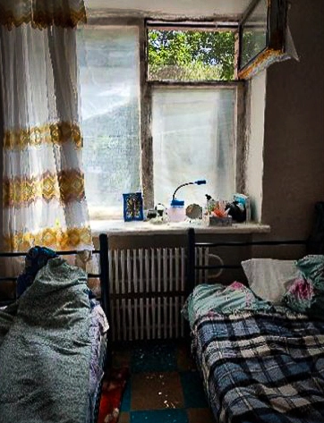 Two narrow beds in a poorly lit room with cracked windows and worn-out bedding, showing the difficult living conditions in a collective centre.