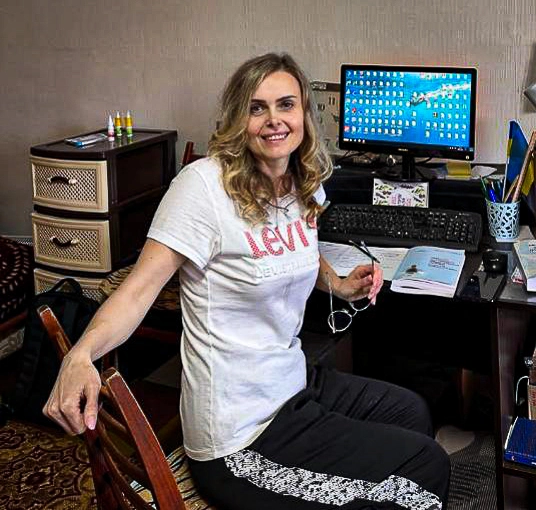 Olena sitting at a desk in her new apartment with a computer, notebooks, and a relaxed smile, representing comfort and stability. 