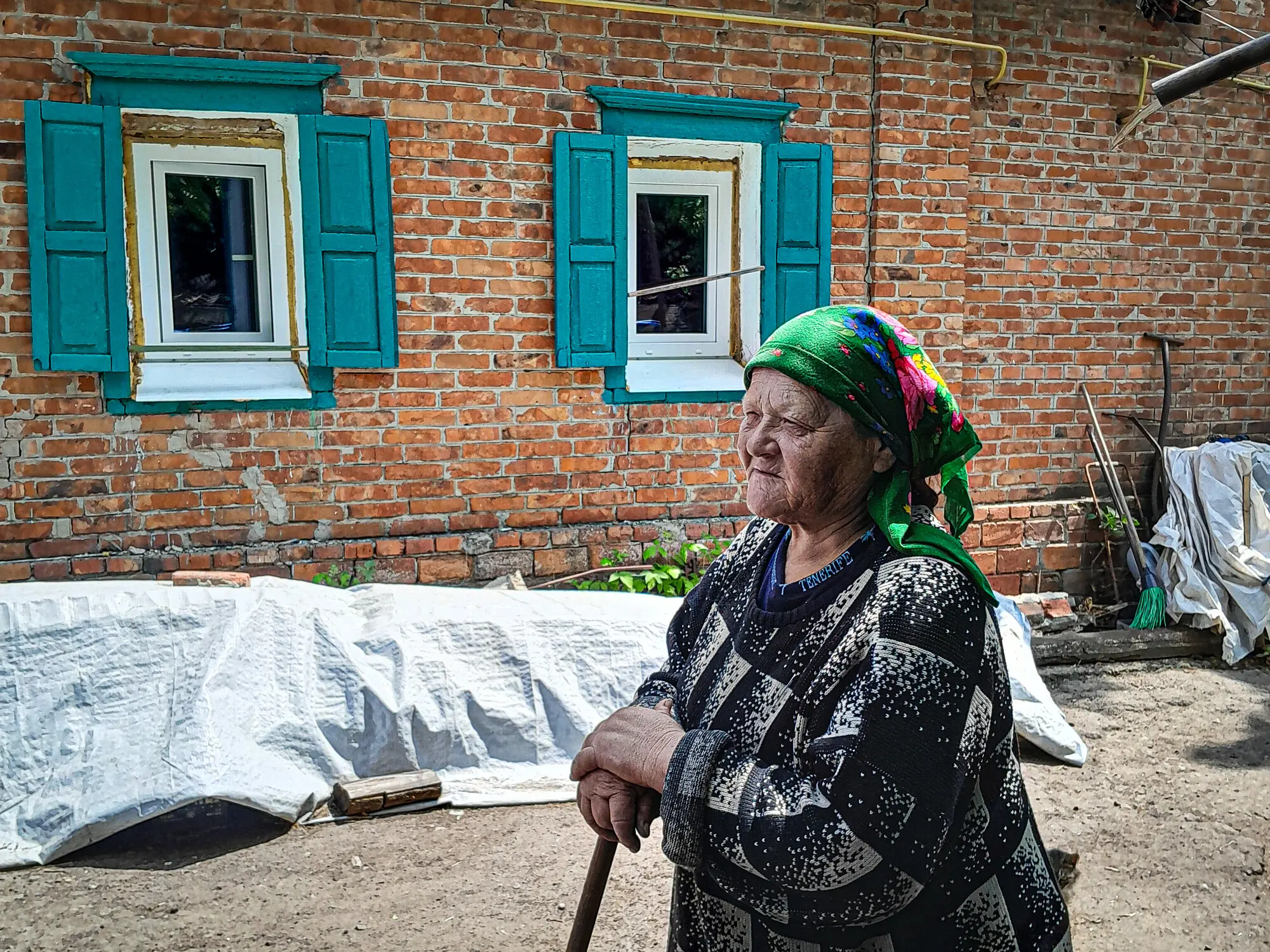 Elderly woman wearing a green headscarf stands outside a brick house with new white-framed windows and turquoise shutters.