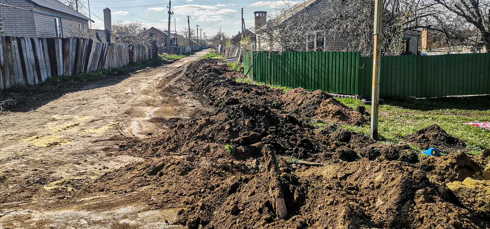 Dirt road with trenches prepared for pipeline installation beside village homes.