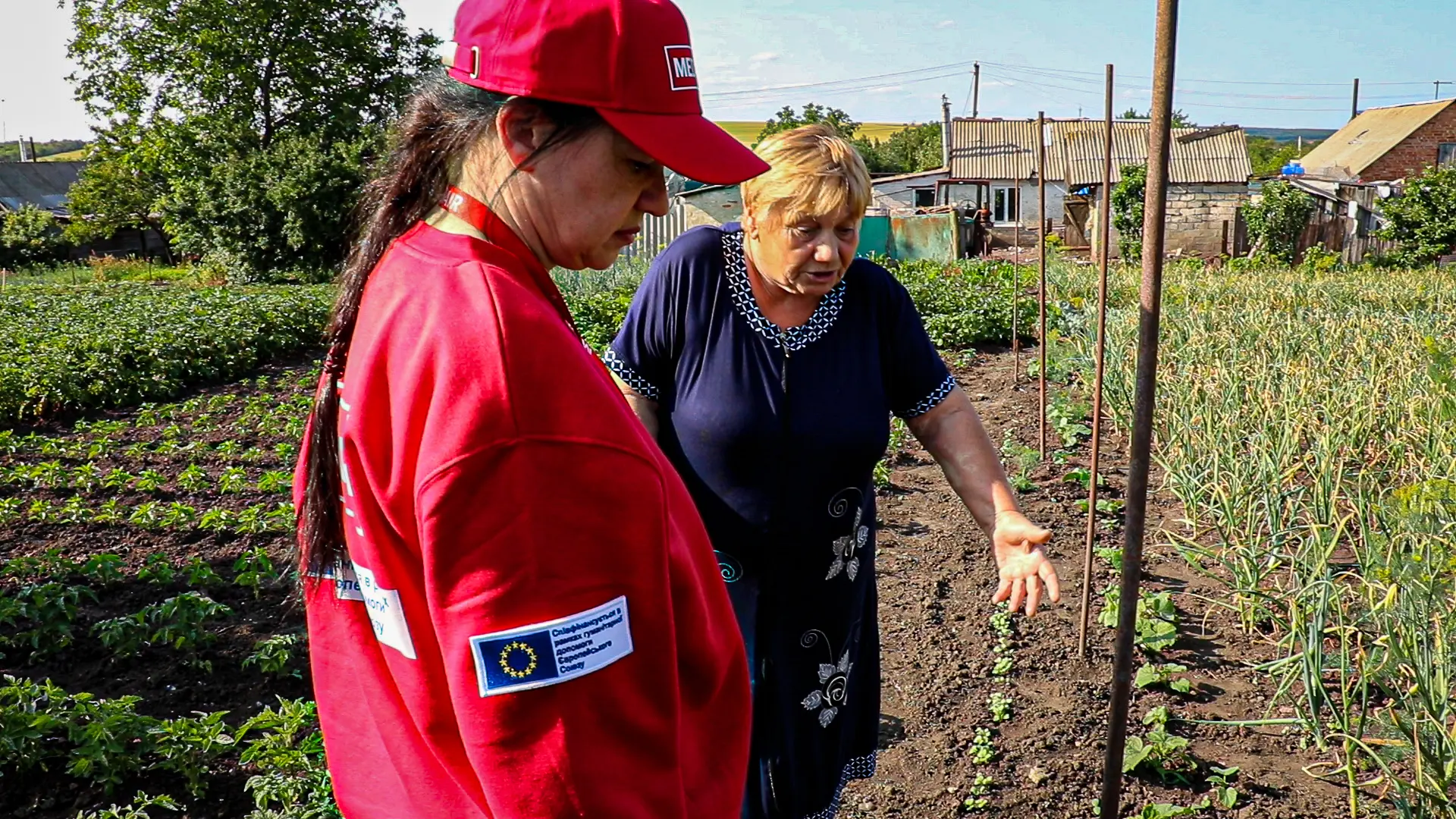 Woman in red jacket listens as an elderly woman explains water access challenges in a vegetable garden.