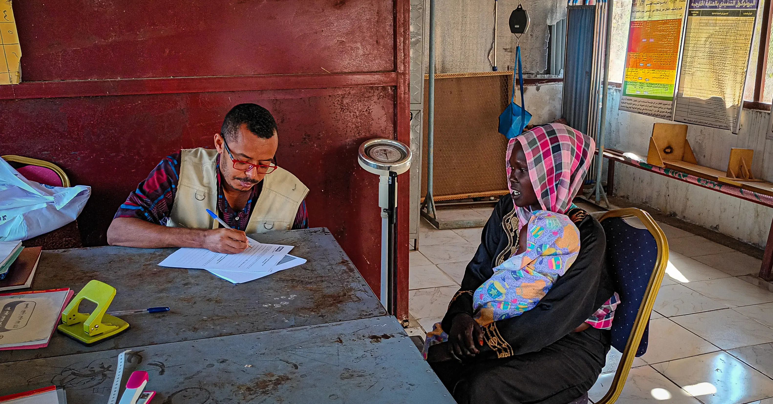 Medair staff member Elfatih writes on a form while Mariam sits across from him holding her baby Yassin at a health facility in Blue Nile.