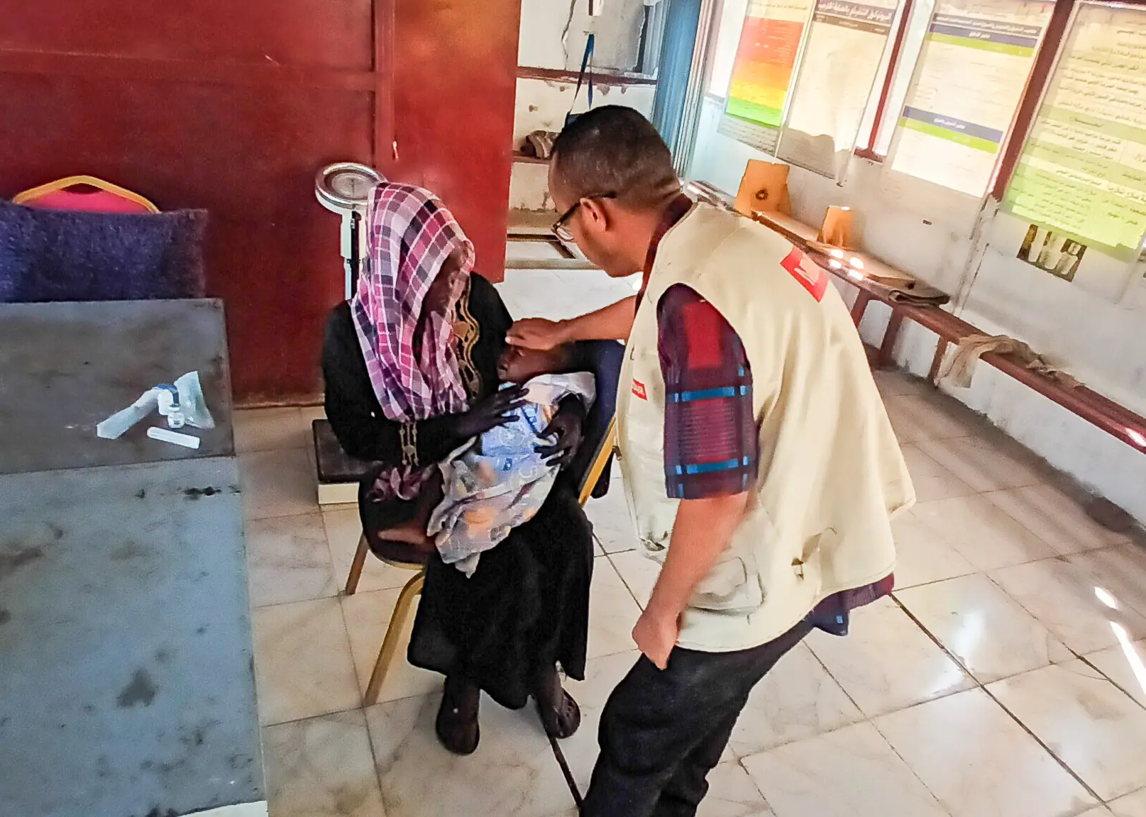 Elfatih leans forward to examine baby Yassin, who is on his mother Mariam’s lap, during a health check-up at a Medair clinic.
