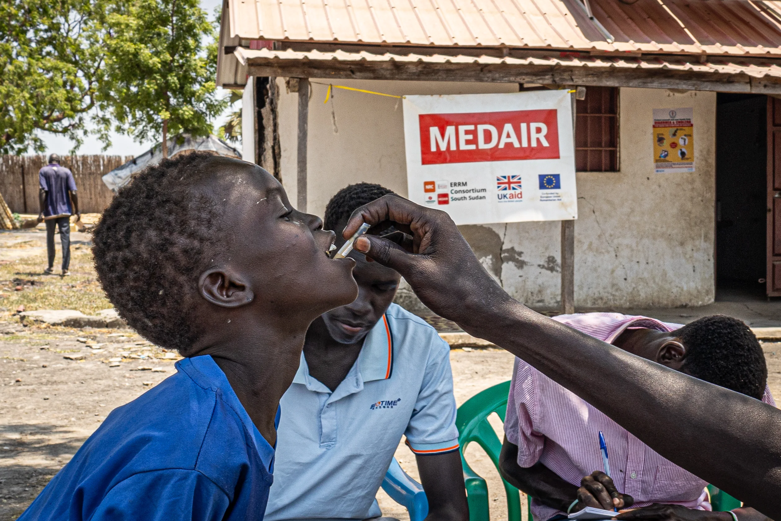  A boy opens his mouth as a health worker administers an oral cholera vaccine, with Medair signage behind.