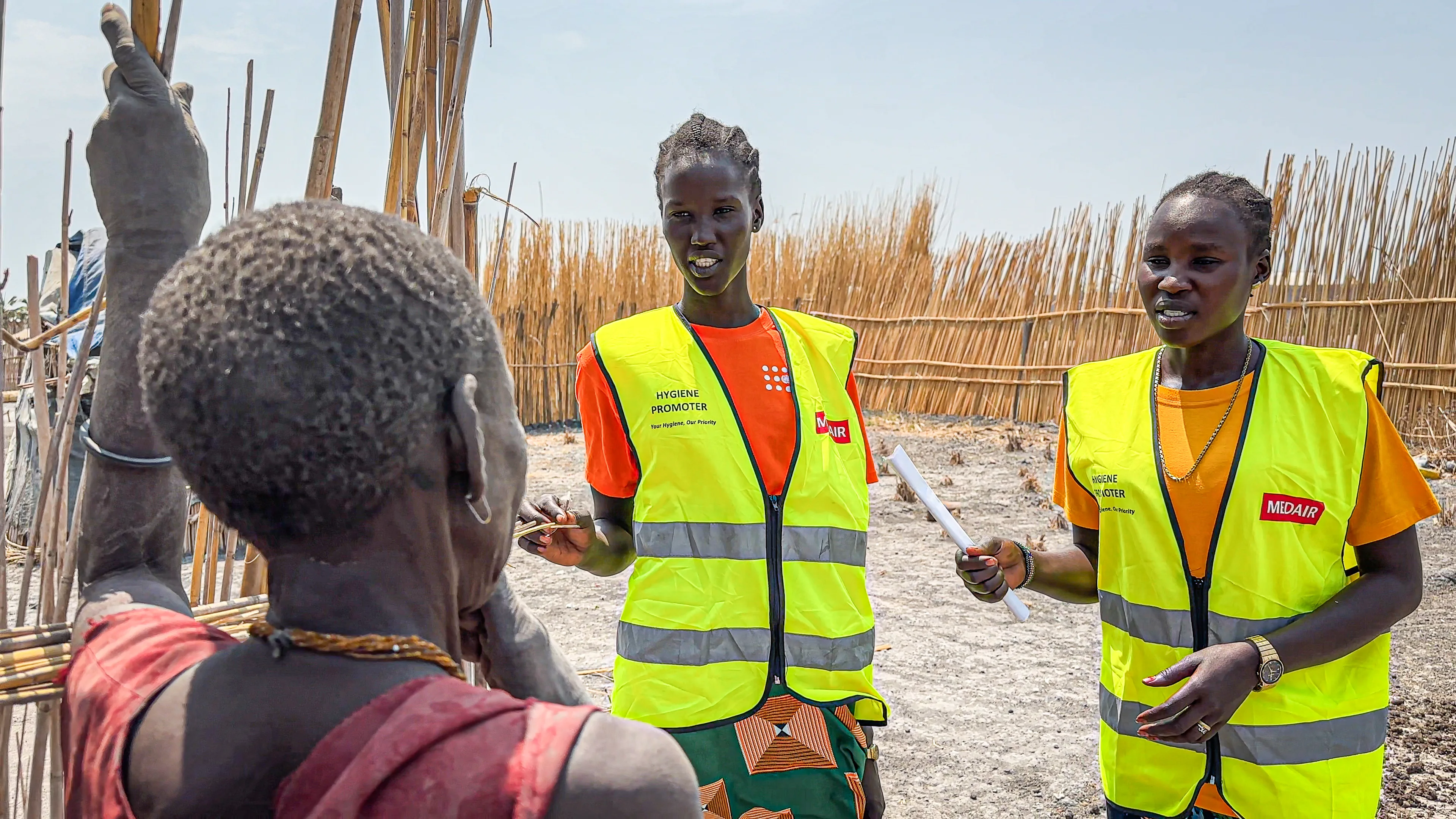  Two Medair hygiene promoters in reflective vests speak to a community member near thatch fencing outside a village.