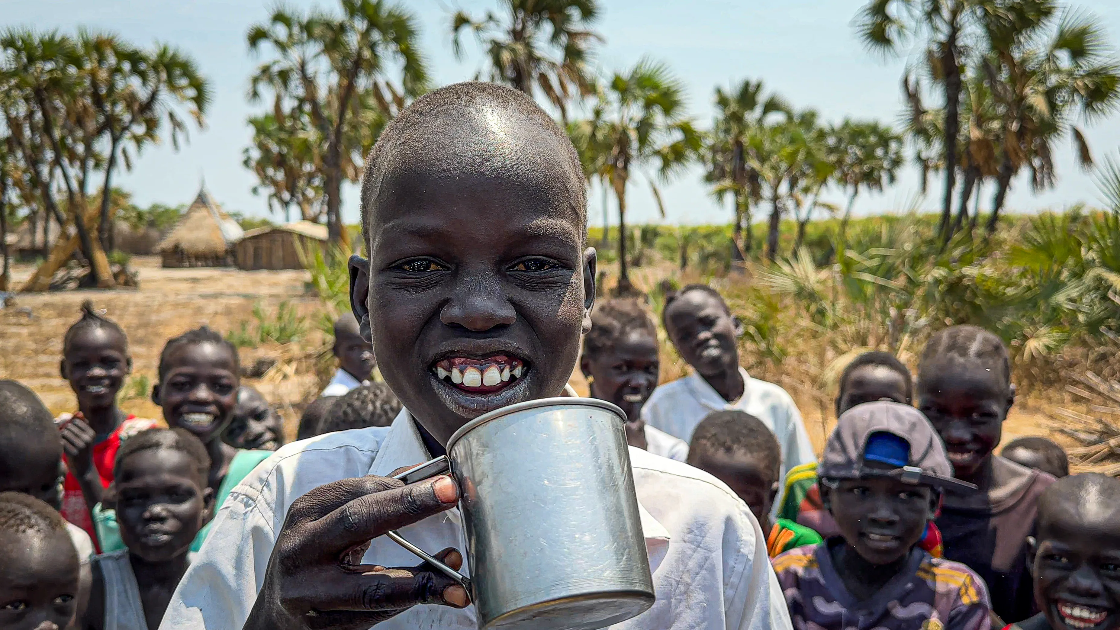 A smiling boy in South Sudan drinks from a metal cup, surrounded by other children and palm trees, highlighting access to clean water after borehole rehabilitation.