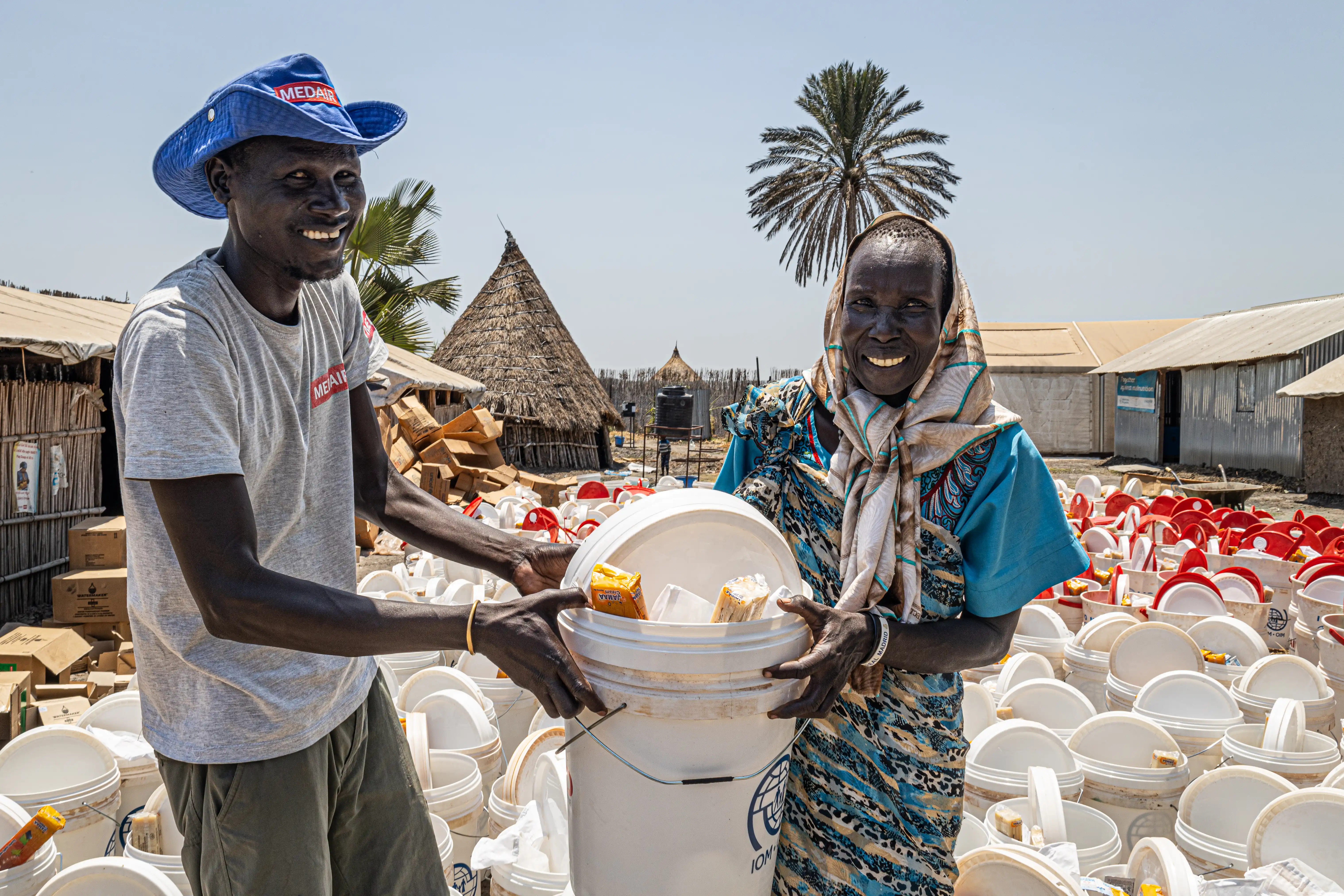 A Medair staff member and a smiling woman hold a bucket filled with hygiene items like soap and chlorine tablets, during a large-scale hygiene kit distribution in a South Sudanese village.