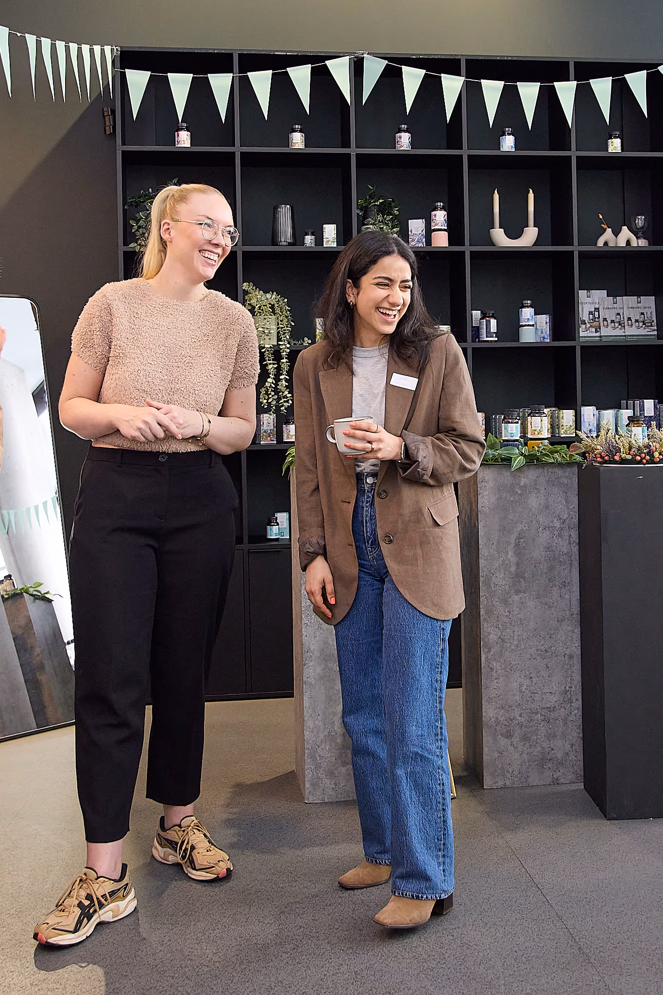 Zwei Frauen lachen, eine hält eine Tasse, im Hintergrund ein Regal mit Produkten und Dekoration.