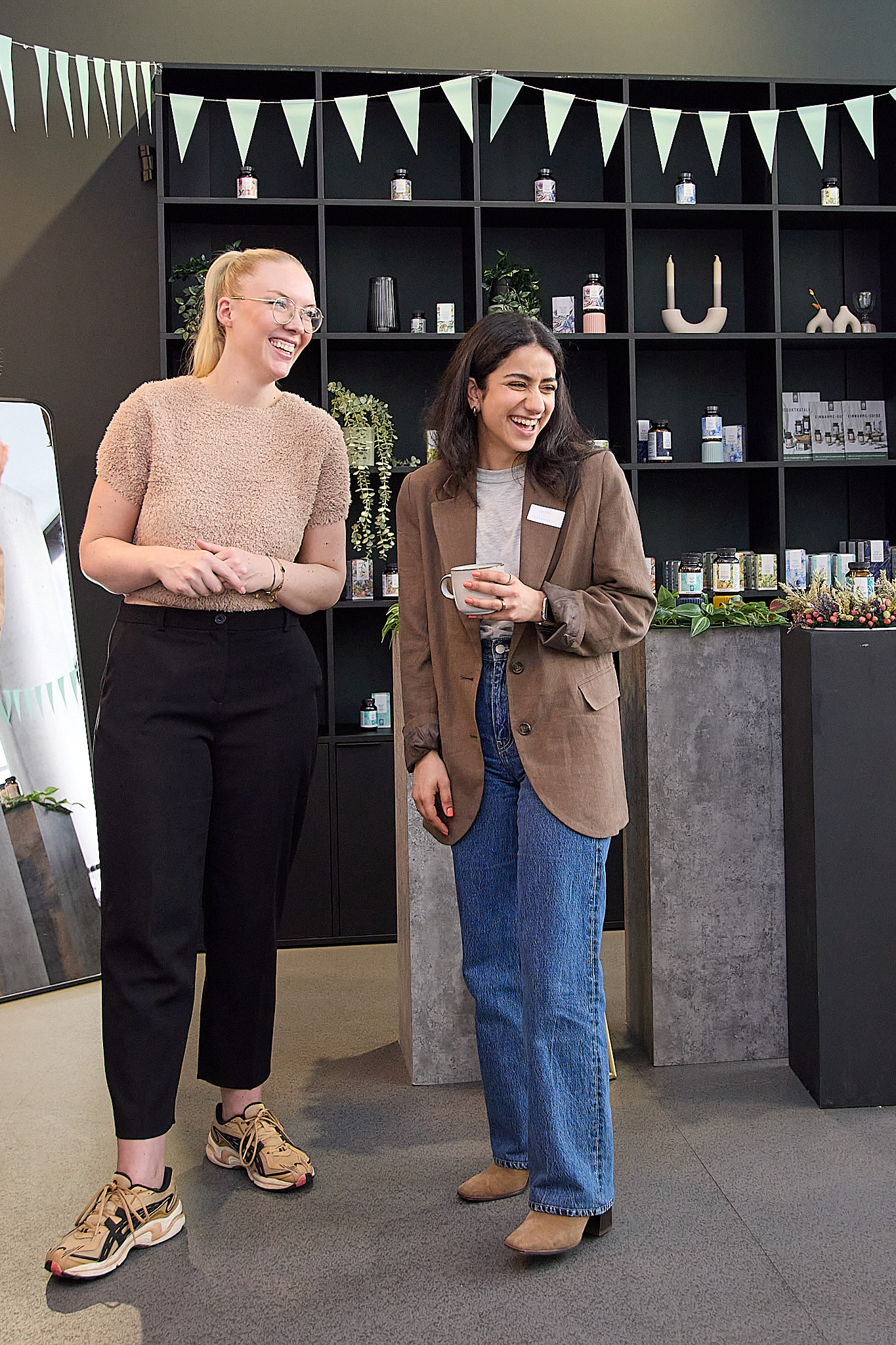 Zwei Frauen lachen, eine hält eine Tasse, im Hintergrund ein Regal mit Produkten und Dekoration.