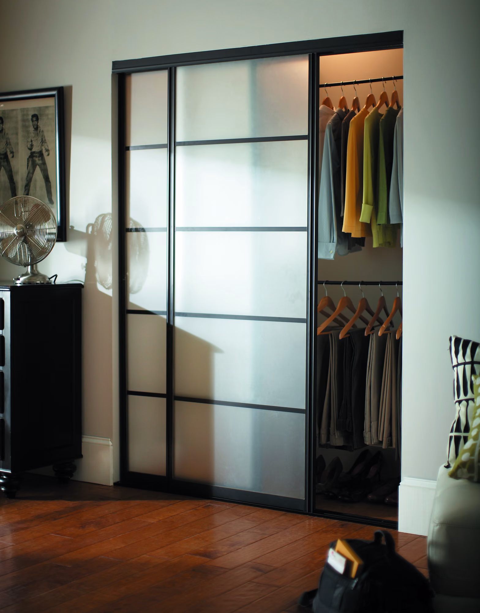 Black-framed frosted glass bypass sliding closet doors opening into a bedroom closet with hanging shirts and pants