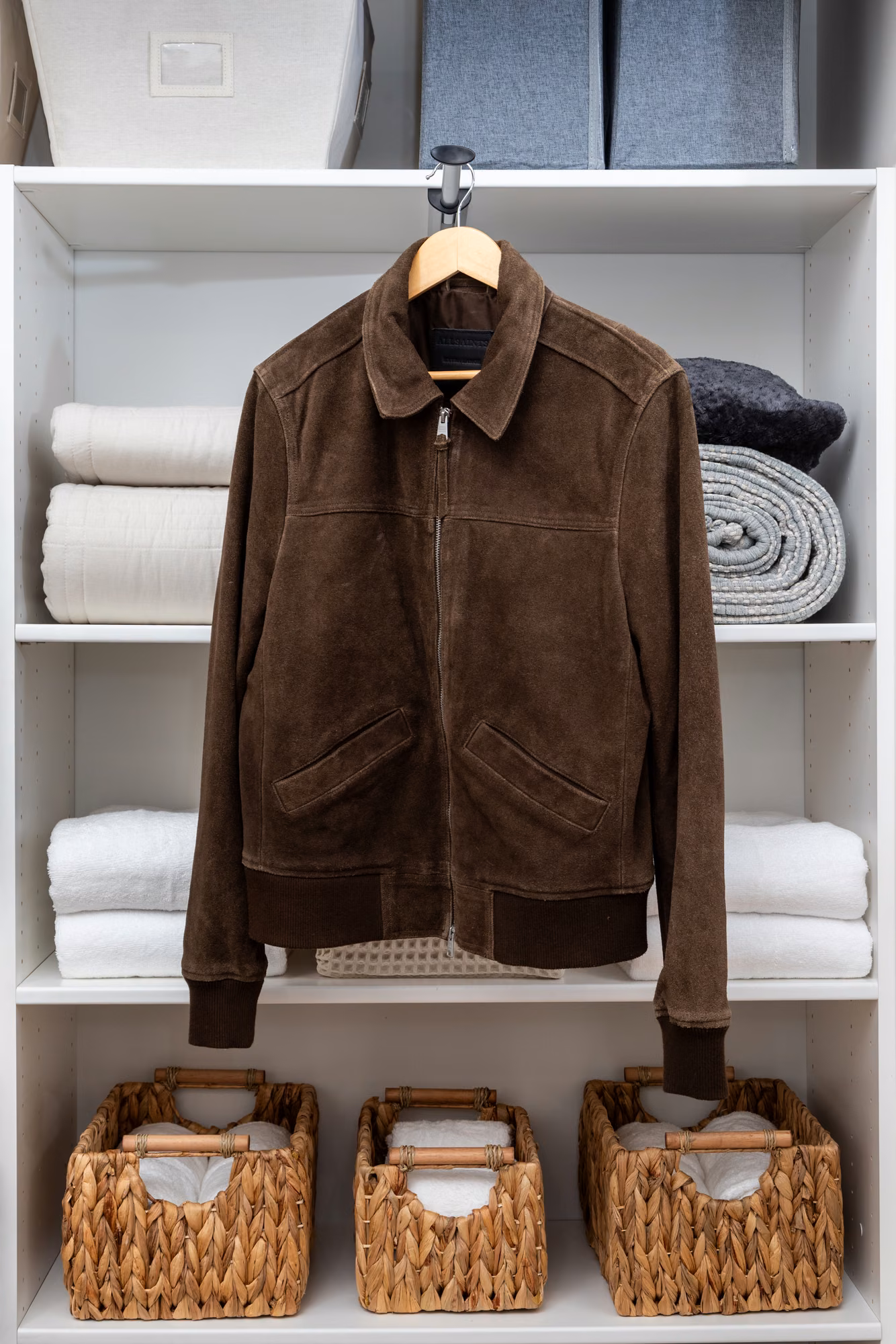 A white linen closet with folded blankets and towels on separate shelves, three woven baskets on the bottom shelf, gray fabric bins on top, and a brown suede jacket hanging from the shelf edge.