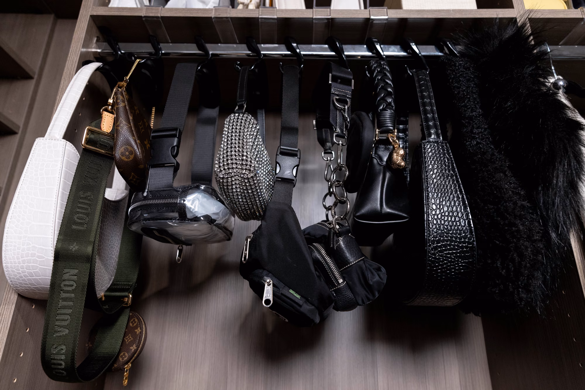A slide-out rack of hooks in a dark wood closet holding about a dozen crossbody bags, belt bags, and shoulder straps.