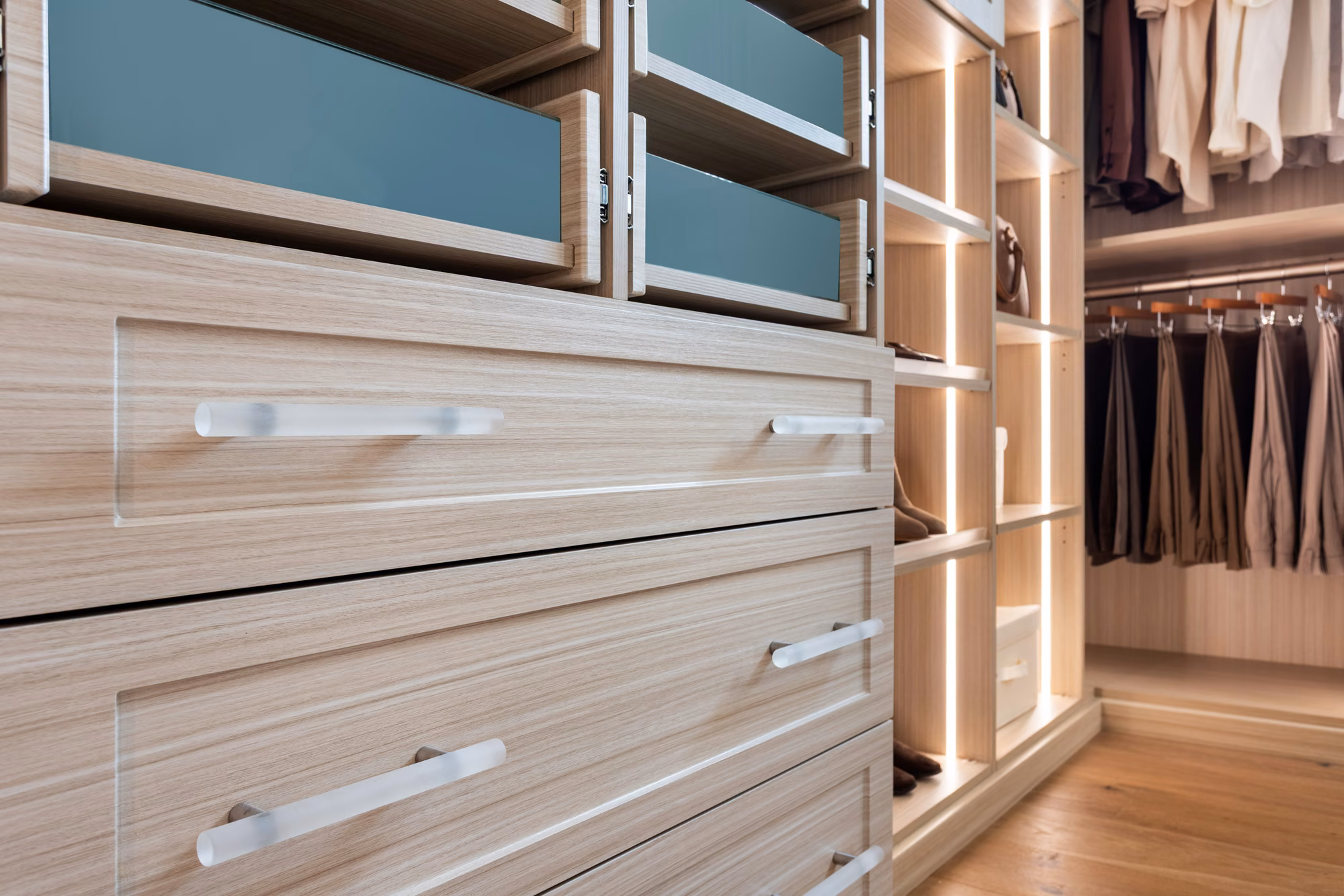 View along a light wood closet showing a bank of drawers with frosted glass fronts and vertical LED strips lighting the shelf columns and hanging sections beyond.