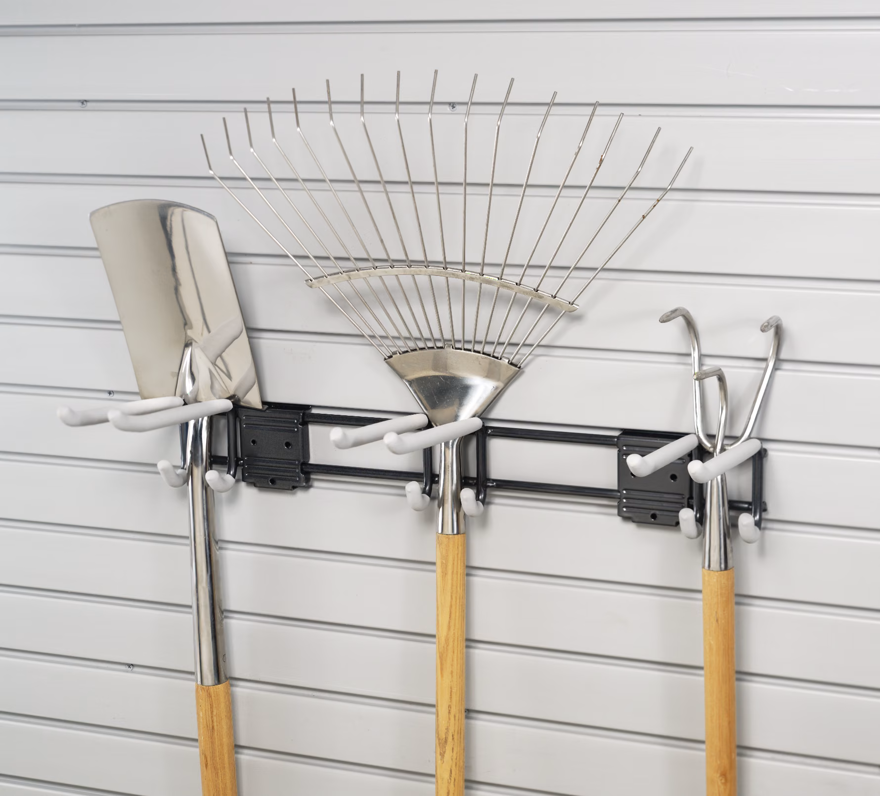 Slatwall rack holding a rake, shovel, and push broom upright against the wall.