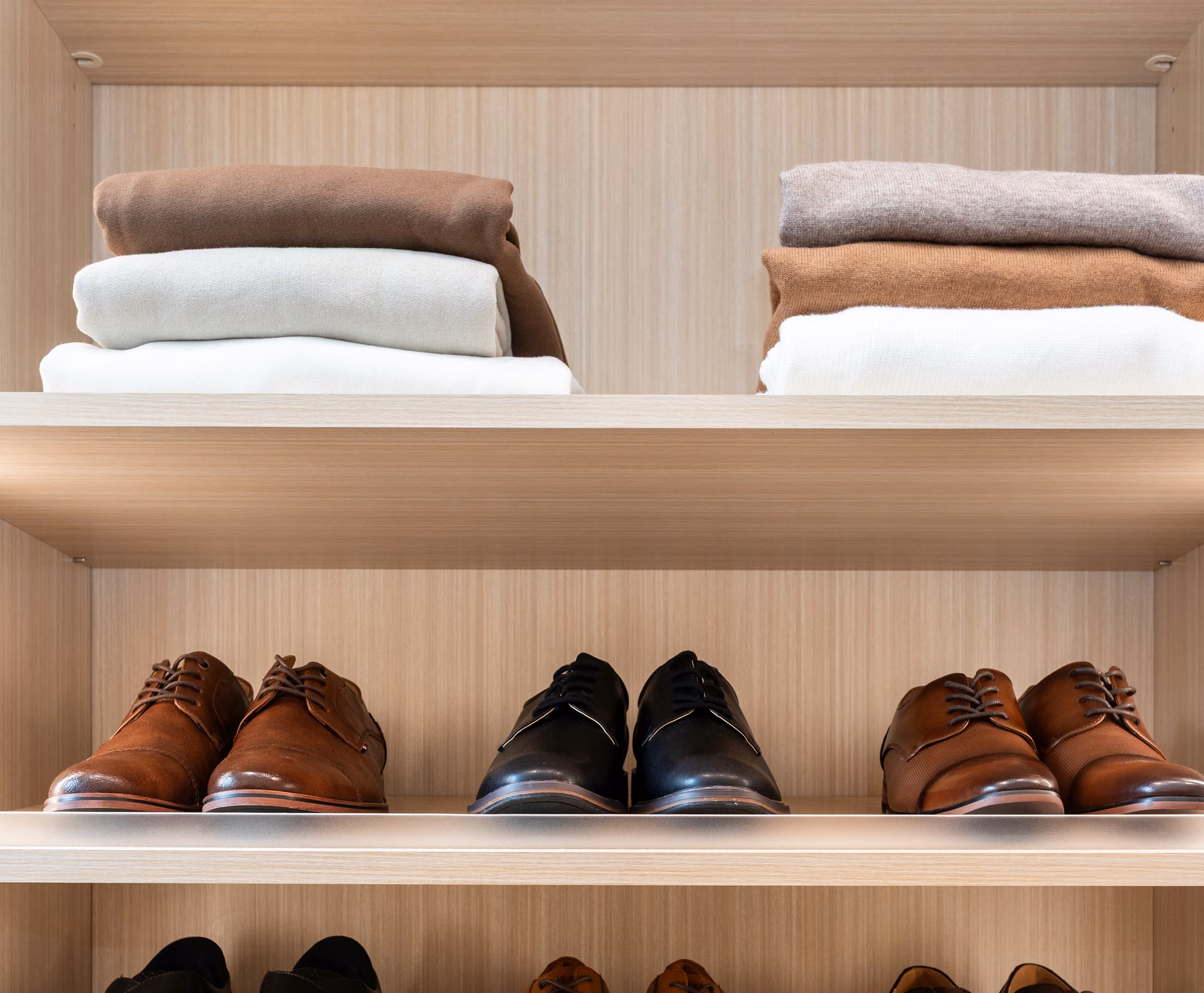 Two stacks of folded sweaters in cream, brown and camel on a lit open shelf, with rows of men's dress shoes on the lit shelves below.