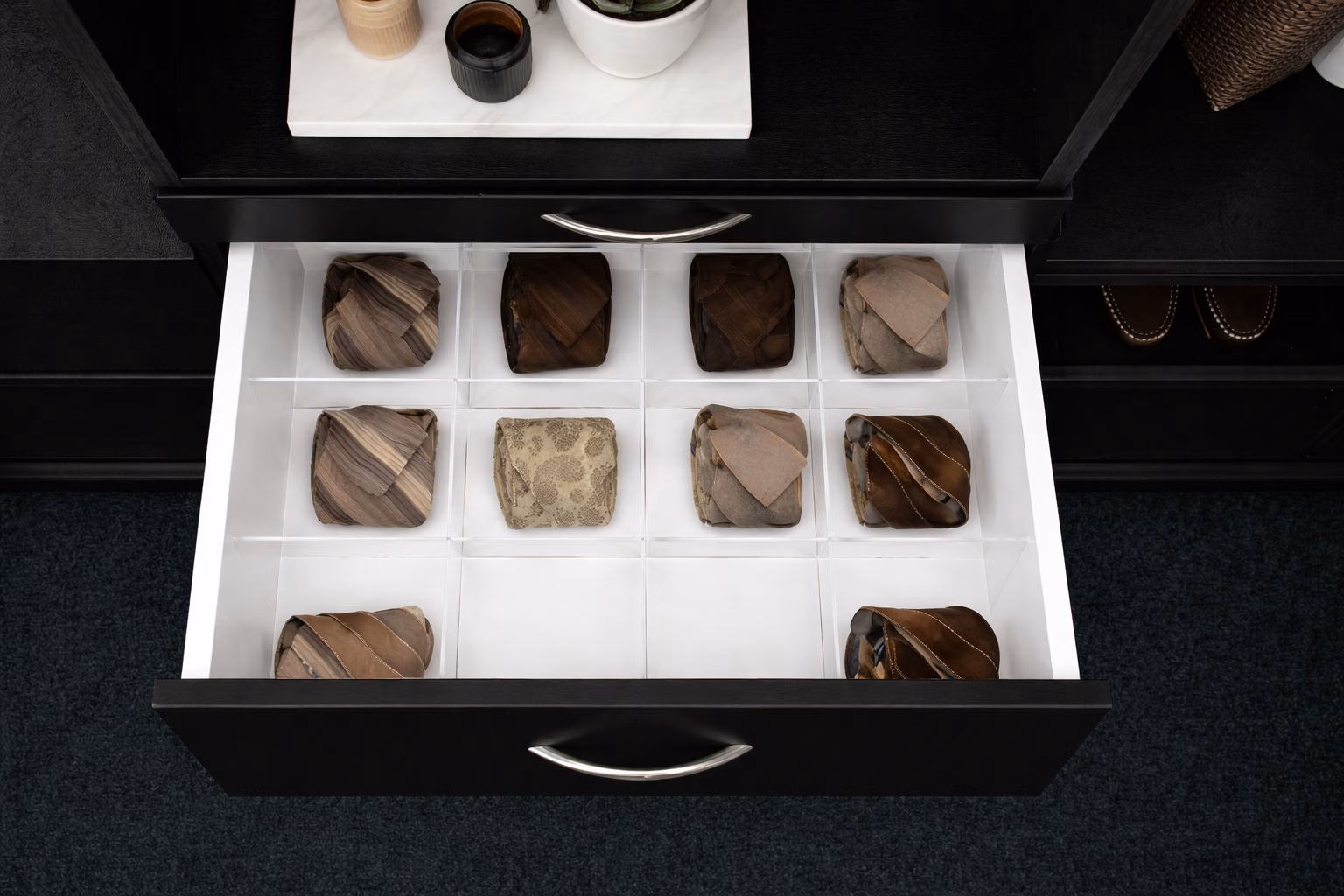 A black closet drawer pulled open to show a clear acrylic grid insert cut to the drawer, forming twelve square compartments with a rolled tie standing in nine of them.