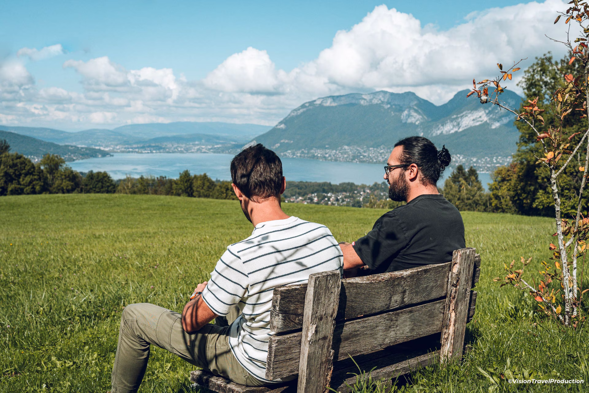 Deux hommes assis sur un banc en bois surplombant un champ verdoyant, avec un lac et des montagnes au loin sous un ciel bleu nuageux.