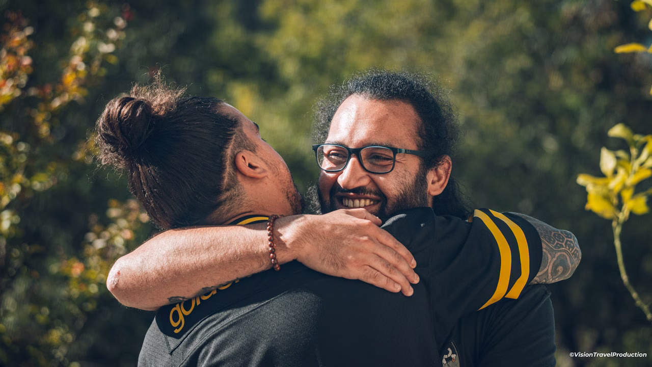 Deux hommes s'enlaçant à l'extérieur, l'un portant des lunettes et souriant largement, sur un fond de verdure.