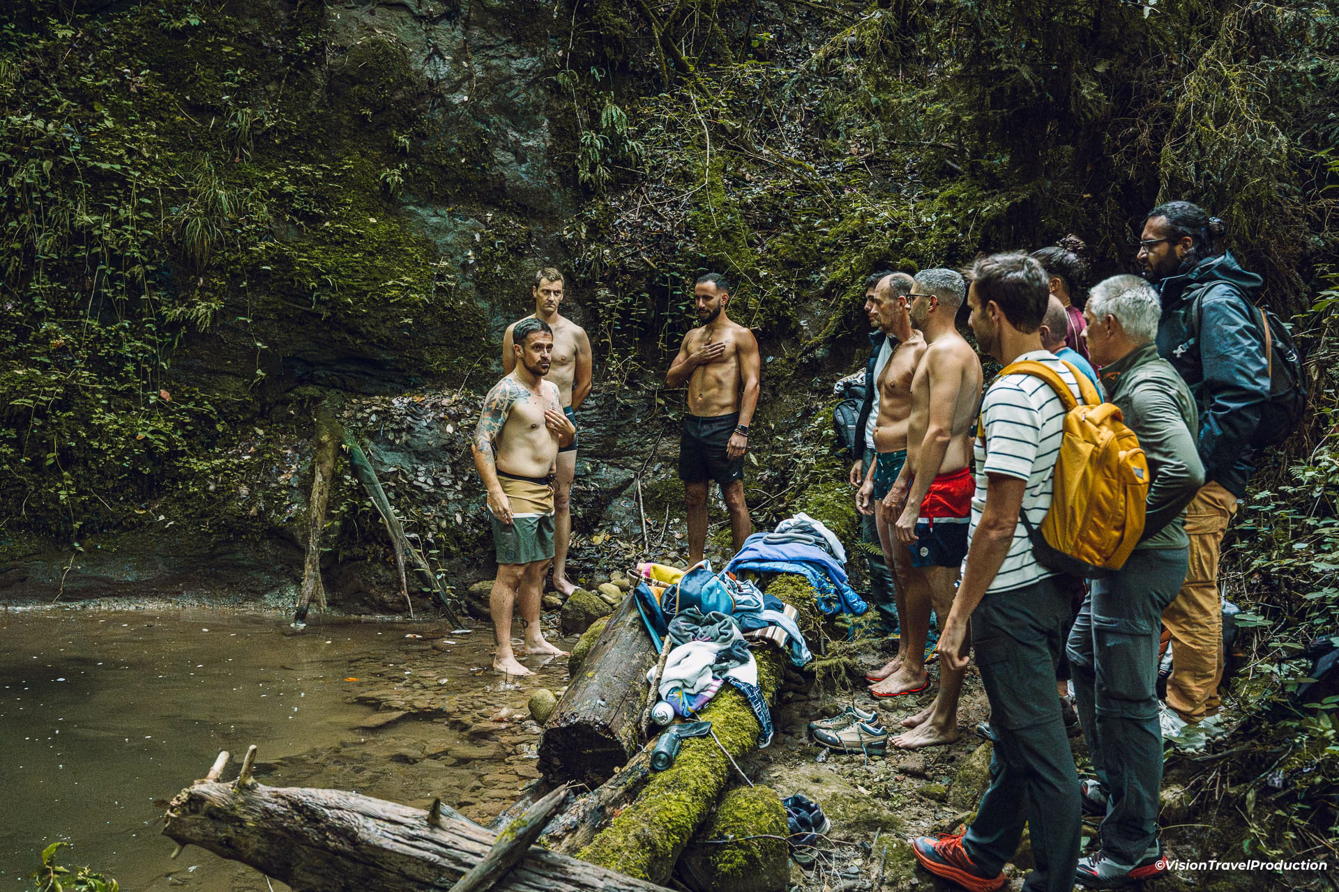 Un groupe d'hommes dans une clairière près d'un point d'eau, certains torse nu et pieds nus, entourant un tronc moussus sur lequel sont posés des vêtements et des chaussures.