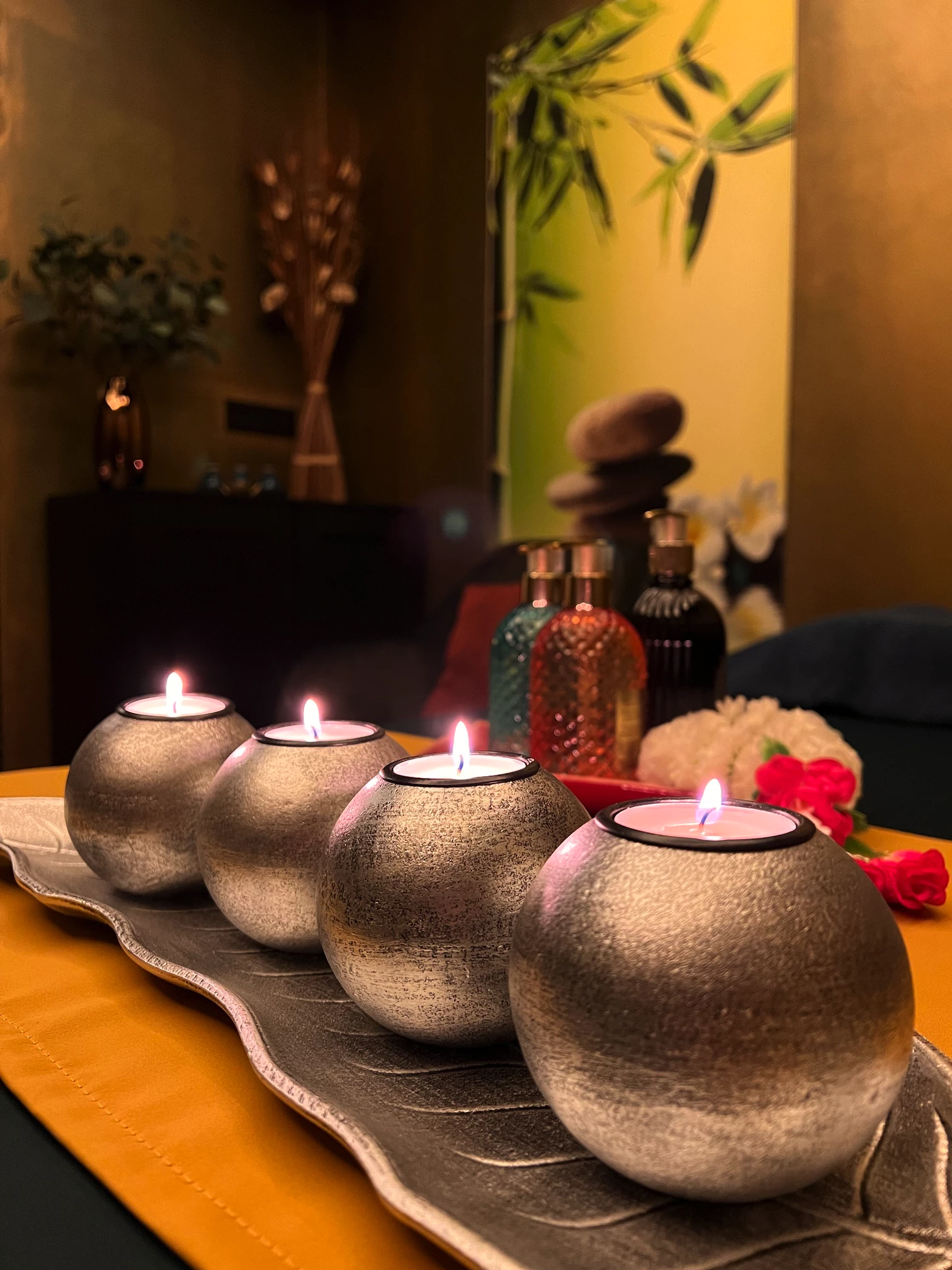 Fotografie pěti stříbrných svíček ve tvaru koule na elegantním stole před vchodem do salonu Touch Massage Center, obklopených různými vonnými oleji a zakřivenými kamennými prvky. Na pozadí je bambusový dekor s měkkým osvětlením, který vytváří klidnou relaxační atmosféru