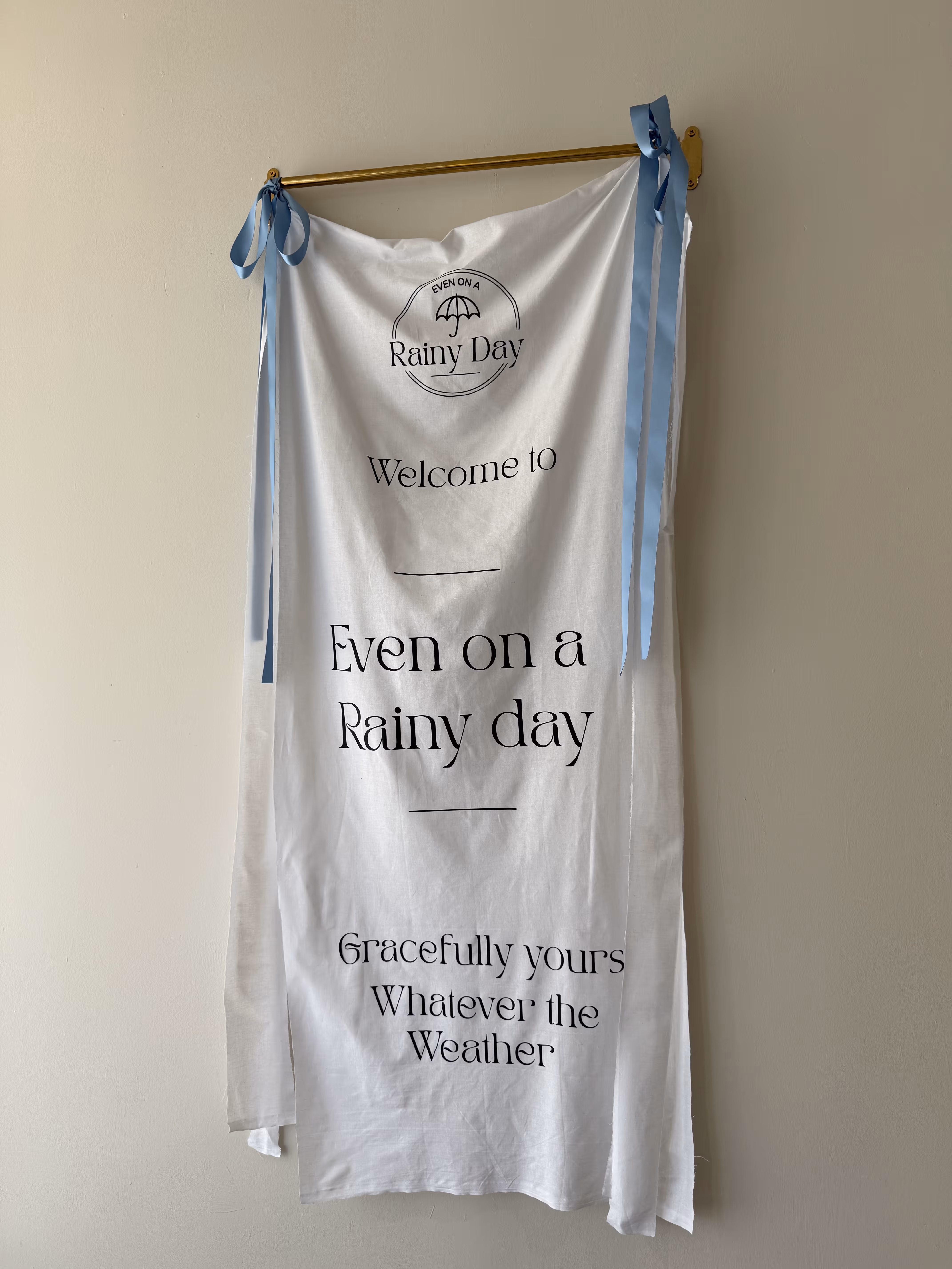 White cloth with blue ribbon ties hanging on a gold rod, featuring the text 'Even on a Rainy Day Welcome to Even on a Rainy day Gracefully yours Whatever the Weather' with a small umbrella graphic.