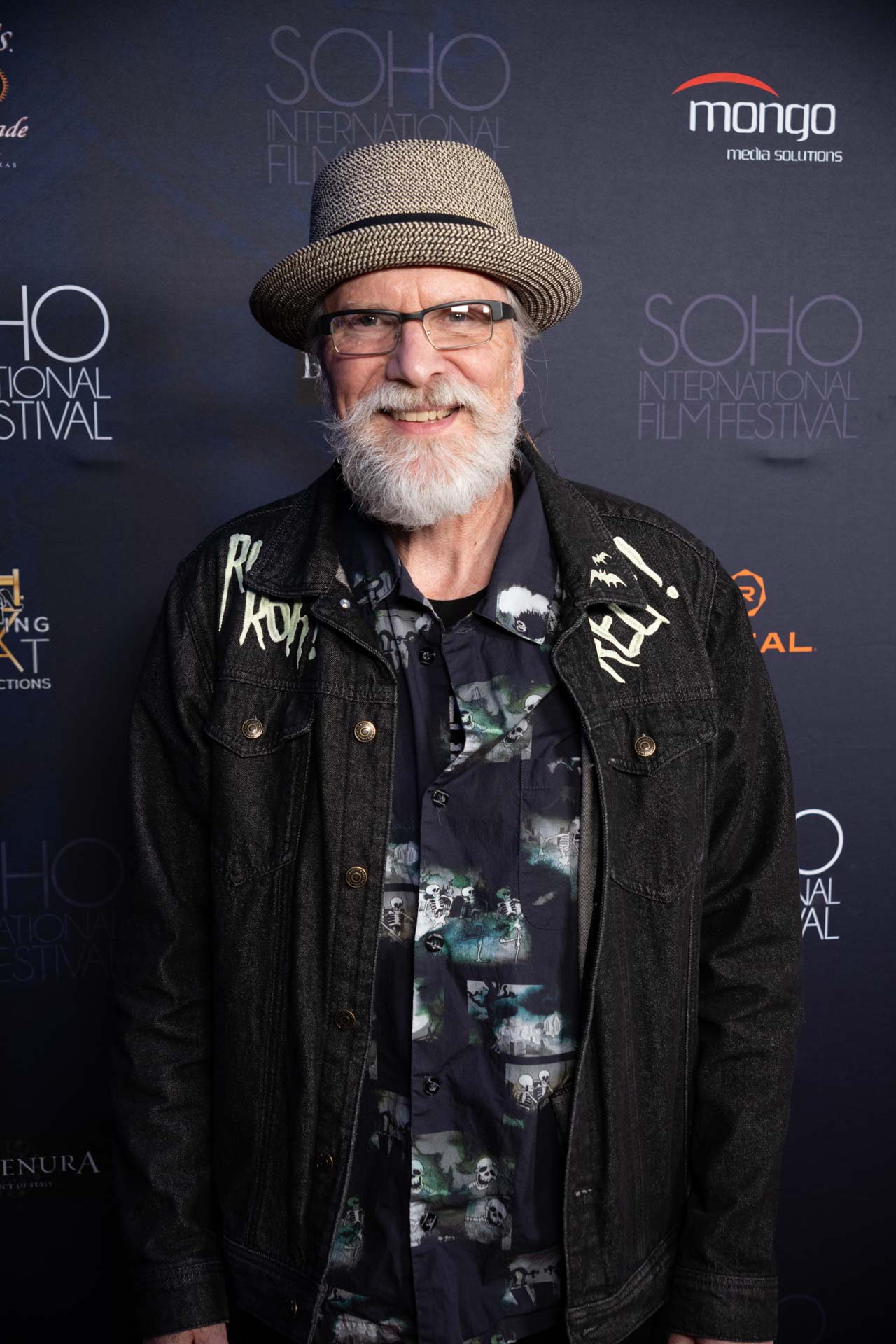Older man with white beard and glasses wearing a patterned shirt, black jacket, and woven fedora hat at the SOHO International Film Festival.
