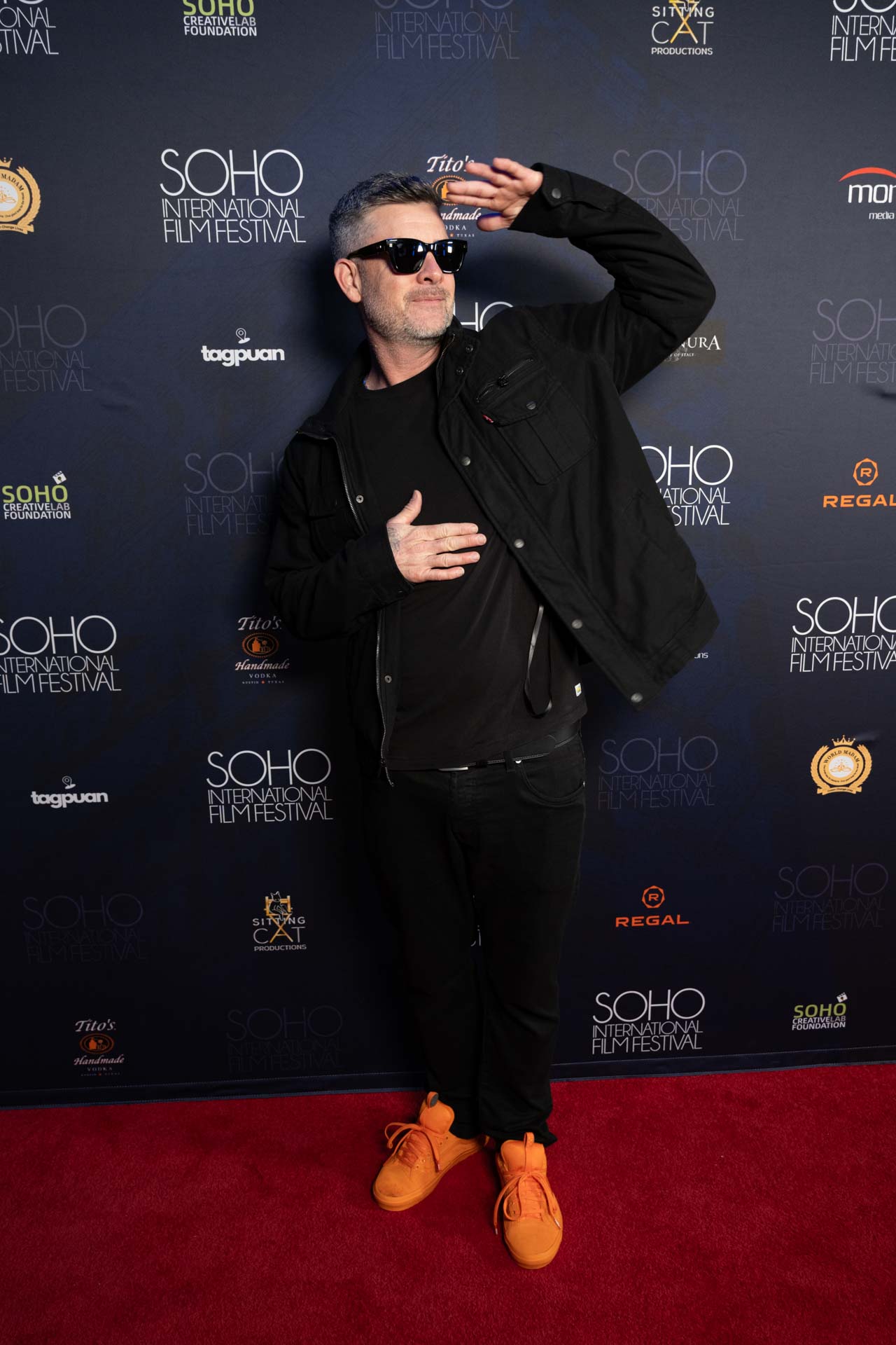 Man wearing sunglasses, black jacket, black pants, and bright orange sneakers posing with one hand on chest and the other raised on a red carpet at the SOHO International Film Festival.