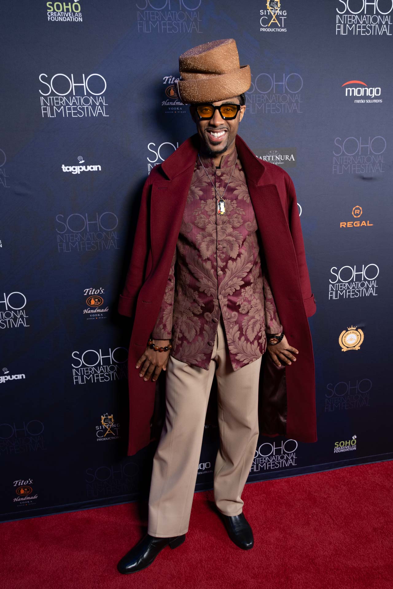 Man smiling on a red carpet wearing a maroon patterned shirt, maroon overcoat draped over shoulders, beige pants, black boots, a textured brown hat, and yellow-tinted sunglasses at the SOHO International Film Festival.