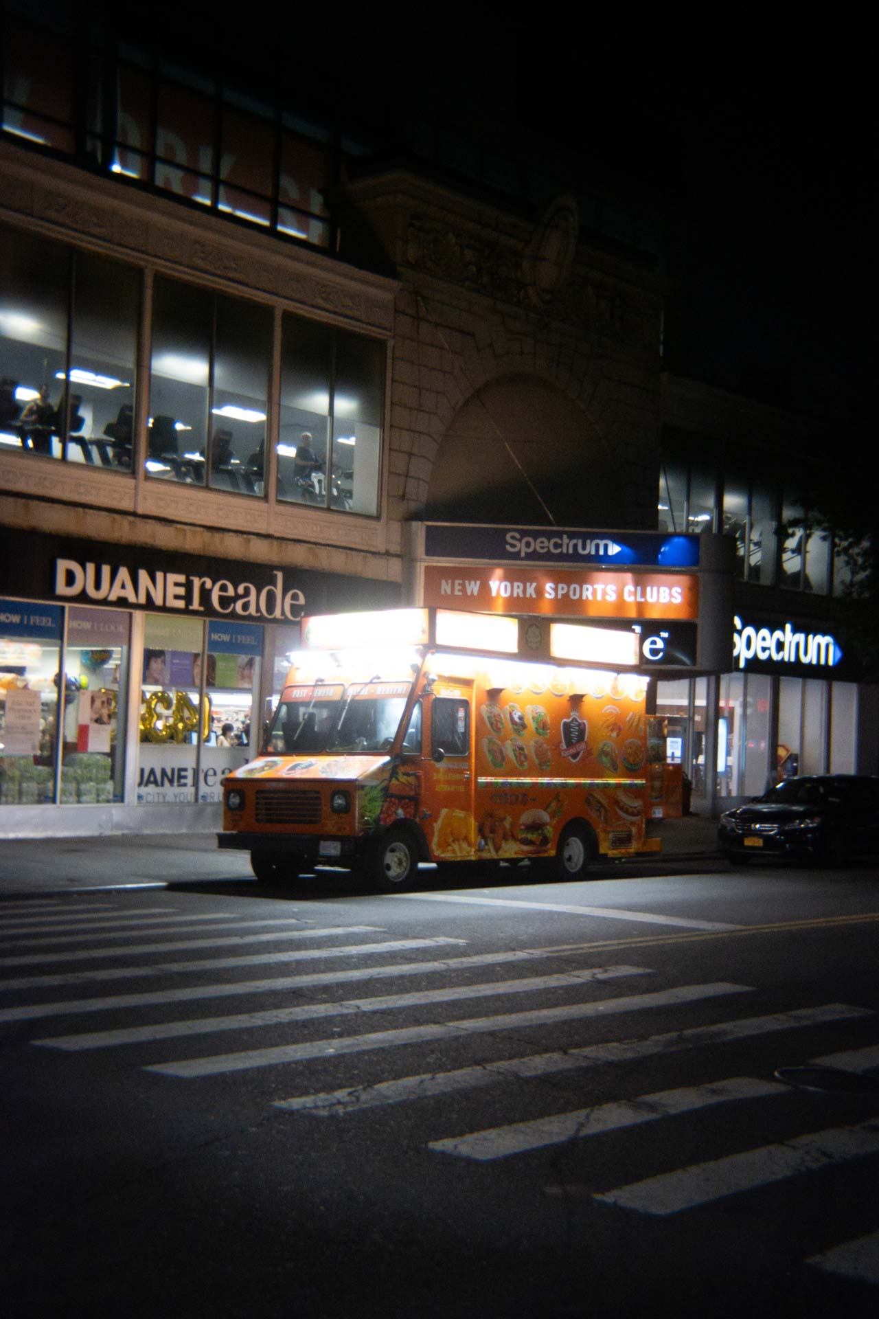 Illuminated food truck with colorful menu parked at a crosswalk on a city street at night in front of Duane Reade and New York Sports Clubs.