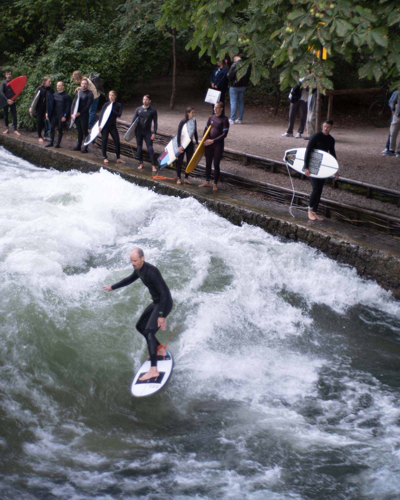 Surfer riding a standing wave in a river with several other surfers in wetsuits holding surfboards on the riverbank.