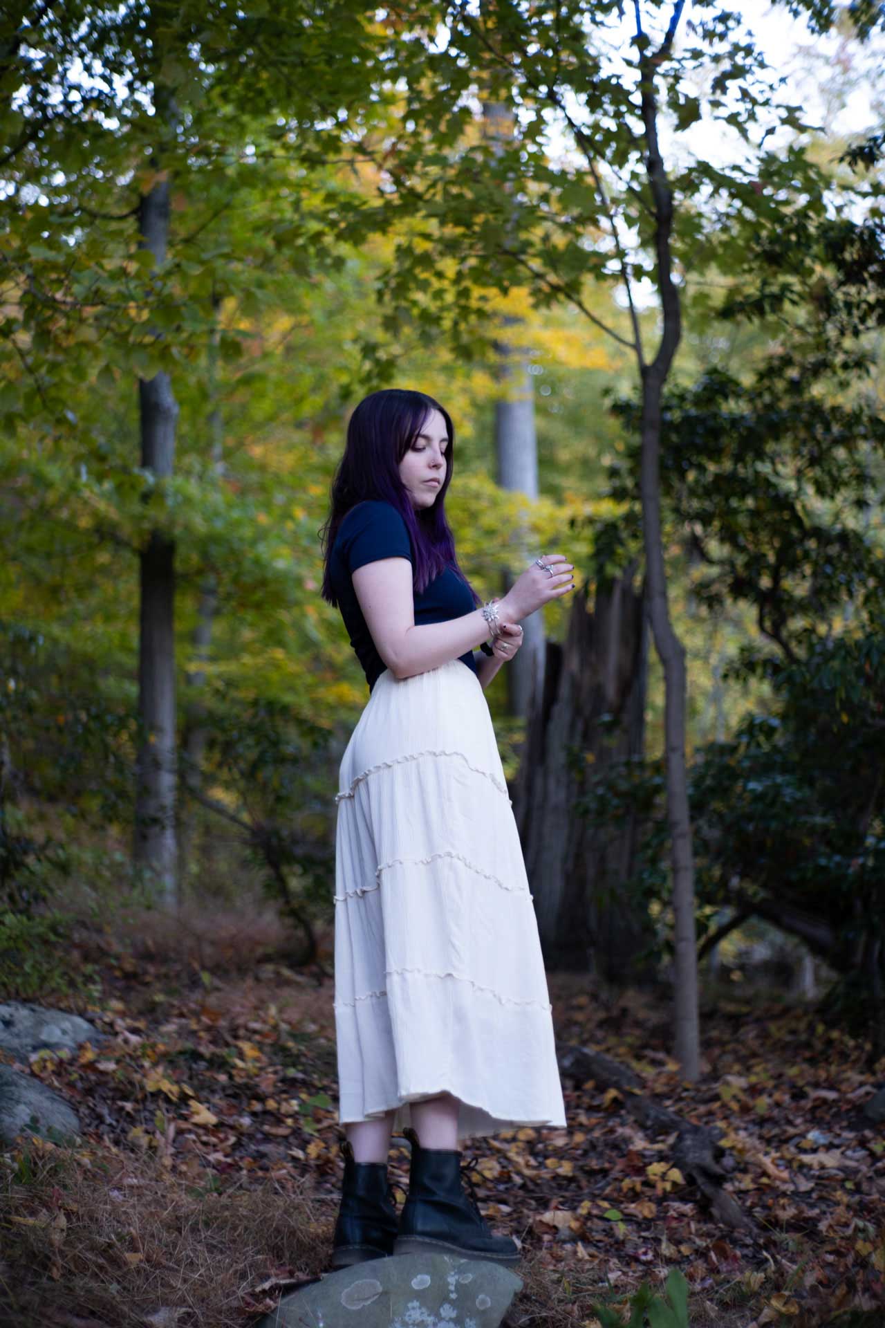 Woman with purple hair wearing a black top, white skirt, and black boots standing on a rock in a forest with autumn leaves.