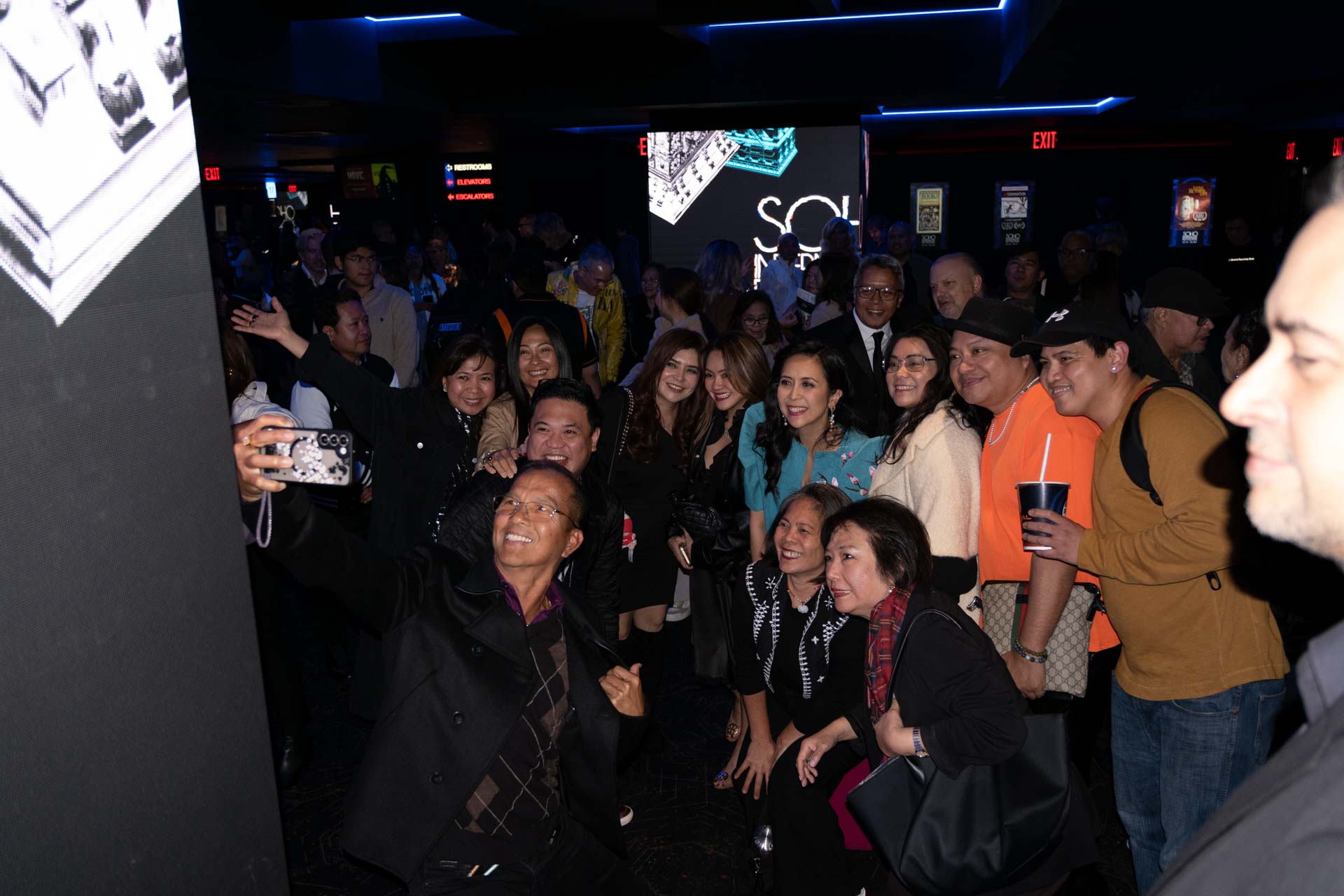 A group of people posing and smiling for a selfie taken by a man in a black coat at an indoor event with dim lighting.