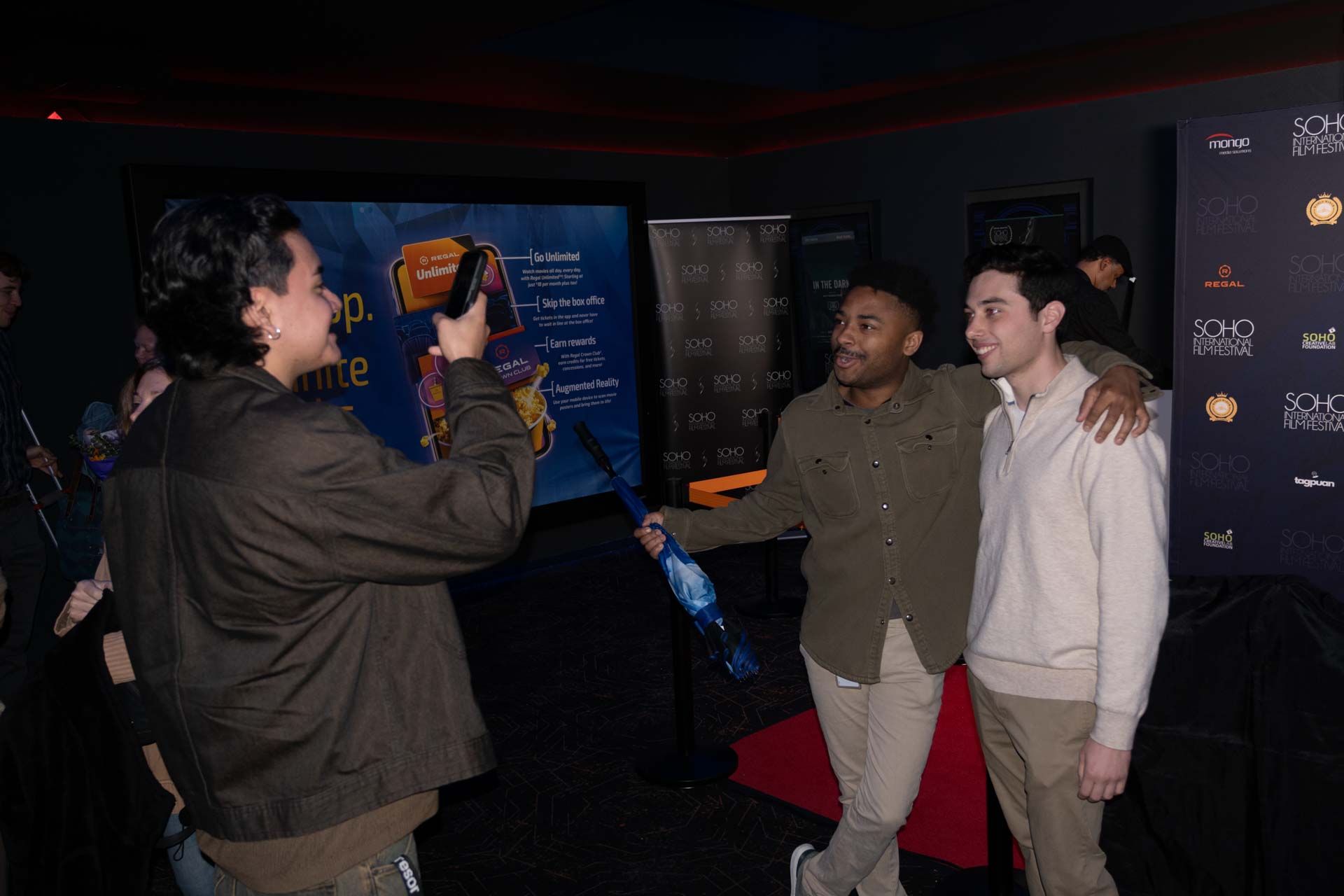 Two men posing happily for a photo taken by a third person holding a smartphone indoors at Soho International Film Festival.