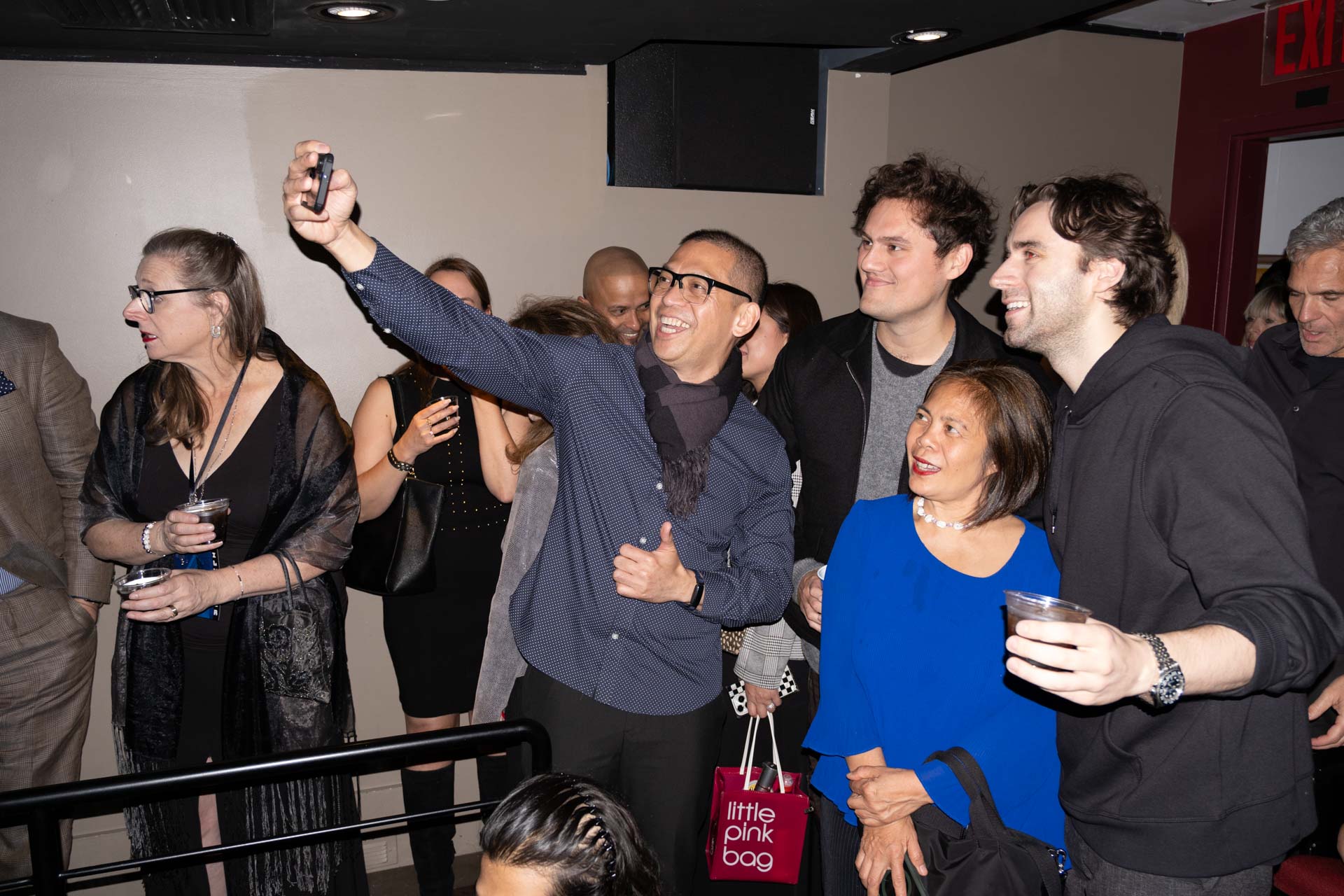 Group of people at an indoor event, one man in glasses takes a selfie with others smiling and holding drinks.