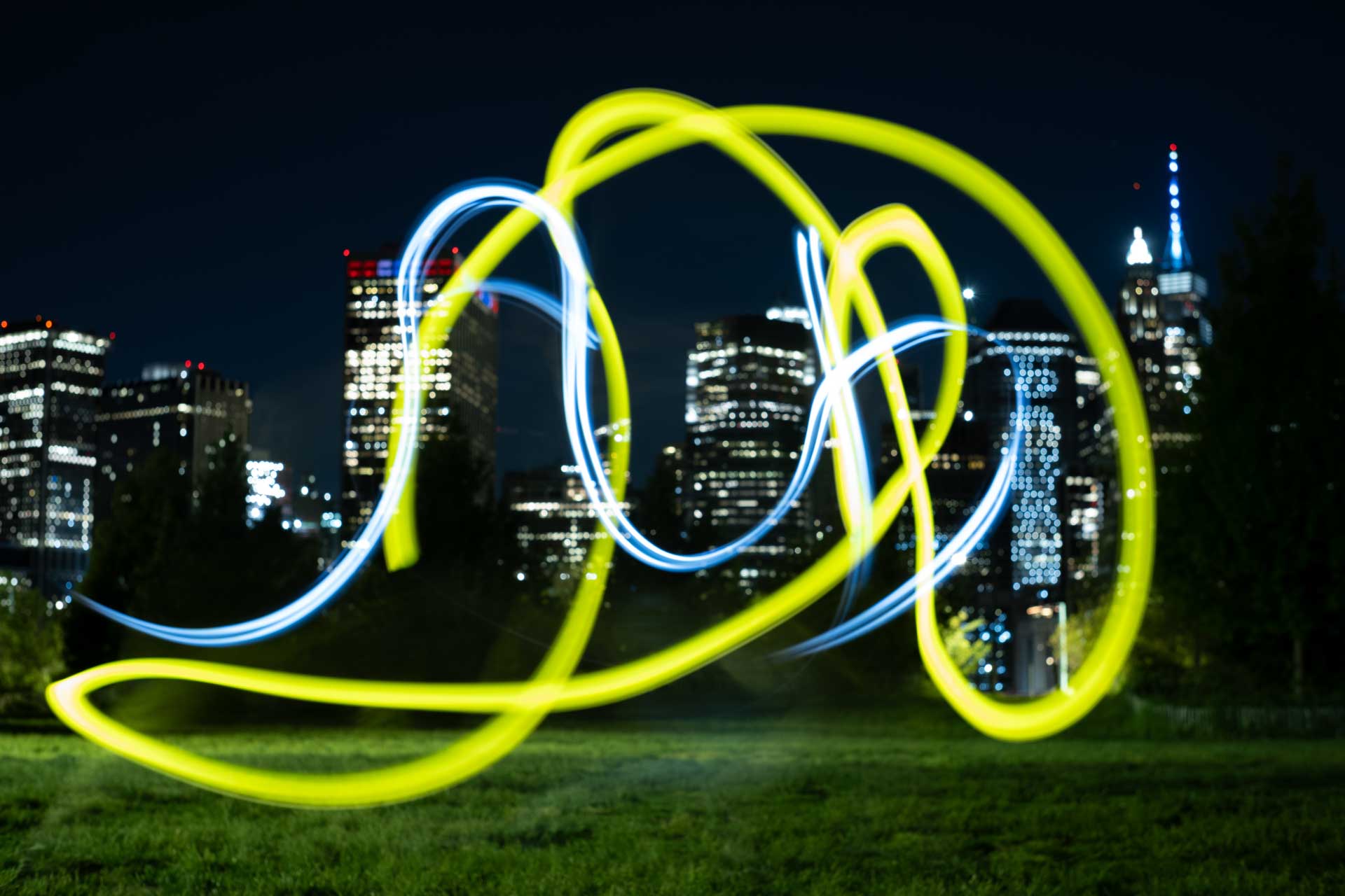 Light trails in yellow and white swirl across a grassy area with a city skyline illuminated at night in the background.