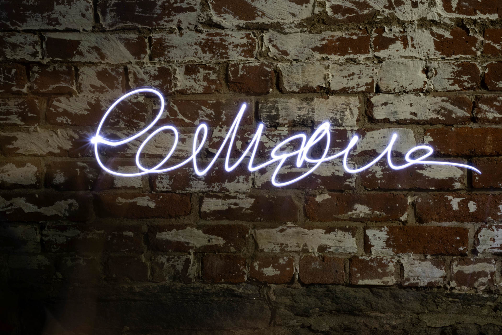 White neon cursive script sign mounted on a brick wall with white paint patches.