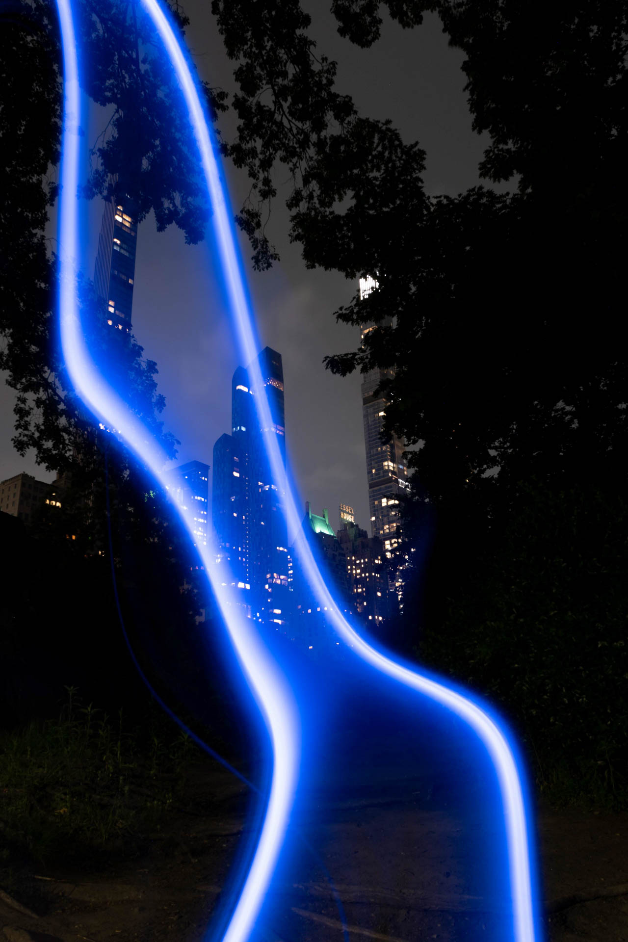 Blue light trails over a dark park path with illuminated city skyscrapers and trees at night.