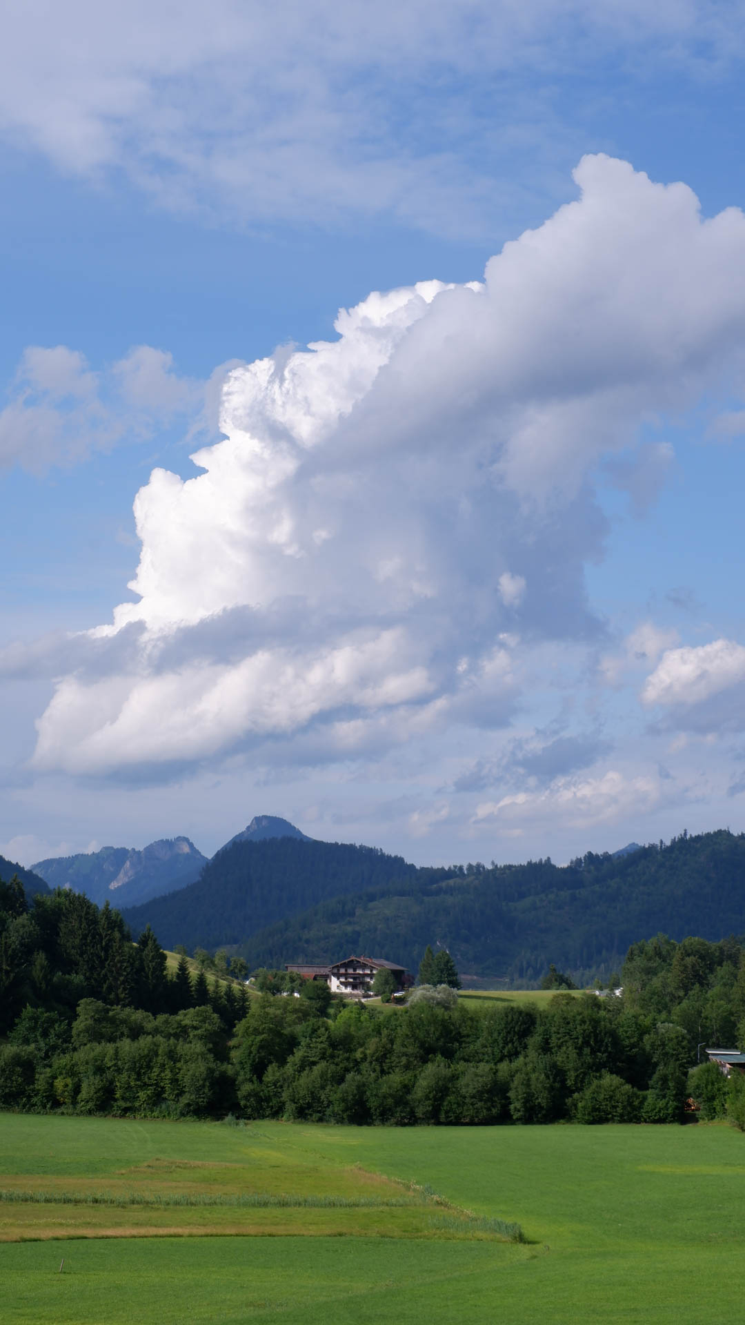 Green fields and forested hills with a house under large white clouds and blue sky.