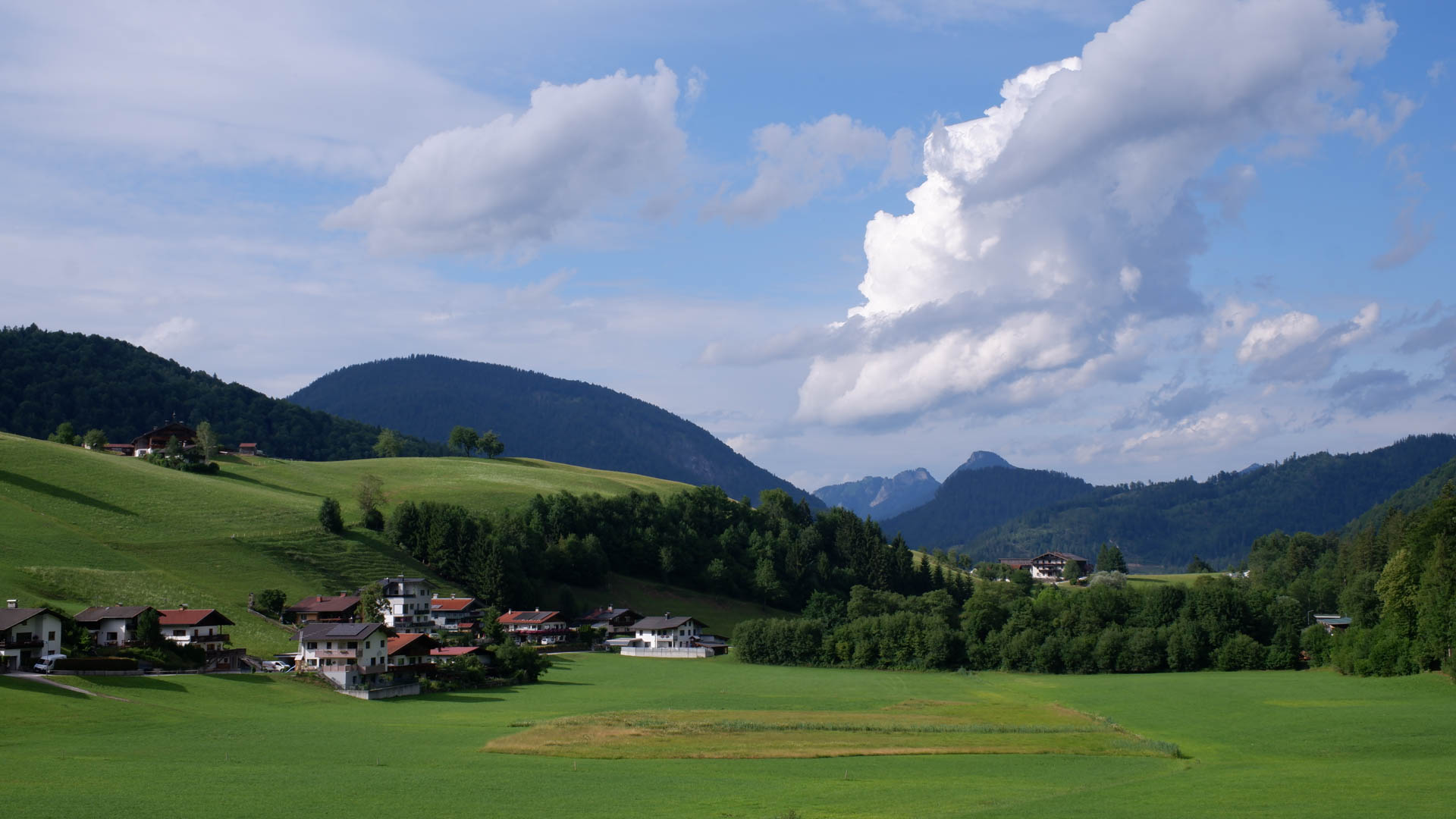 Green fields and houses at the base of forested hills under a partly cloudy sky.