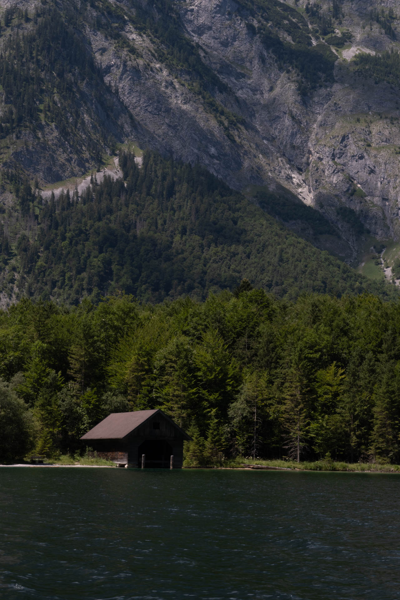 Wooden boathouse on the edge of a forest lake with steep rocky mountains in the background.