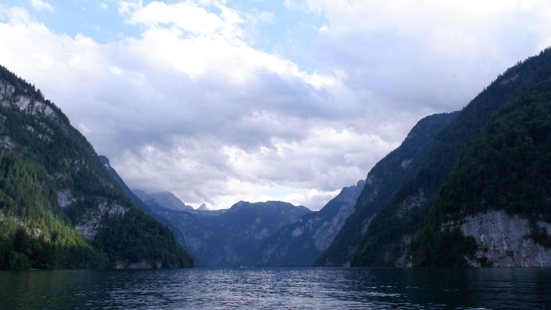 Lake surrounded by forested steep mountains under a partly cloudy sky.