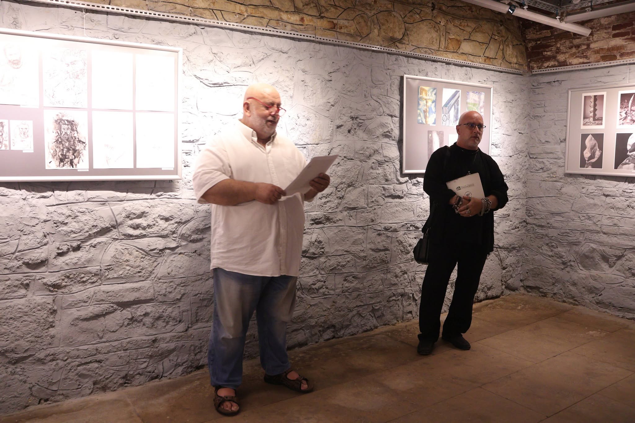 Two people stand in from of a group mail art exhibition. one man presents the exhibition.