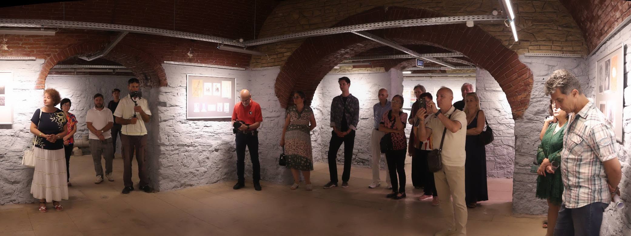 A group of people listen to a presenter introducing a group mail art exhibition.
