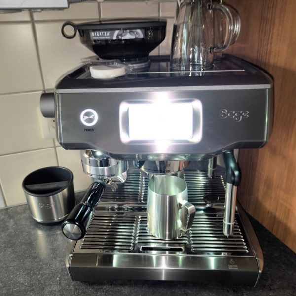 Sage Oracle Touch aparat za espresso sa metalnim bokalom ispod izlaza za kafu i svetlim ekranom, smešten uz drveni kuhinjski zid.