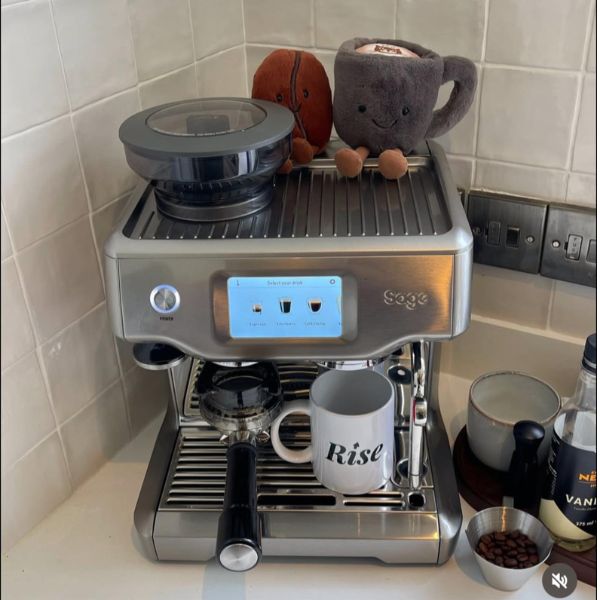 Sage Barista Touch espresso aparat od nerđajućeg čelika na kuhinjskom pultu, sa šoljom ‘Rise’ i plišanim igračkama u obliku šolje i zrna kafe na vrhu.