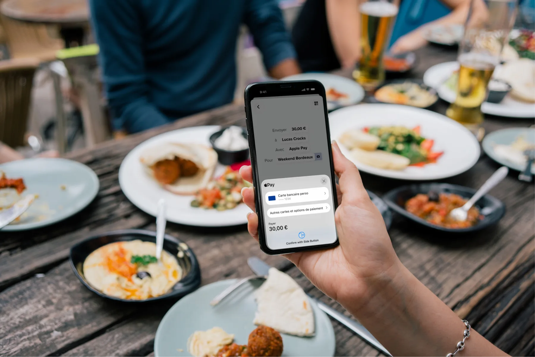 Main tenant un smartphone affichant le paiement Apple Pay de 30 € lors d'un repas en groupe.